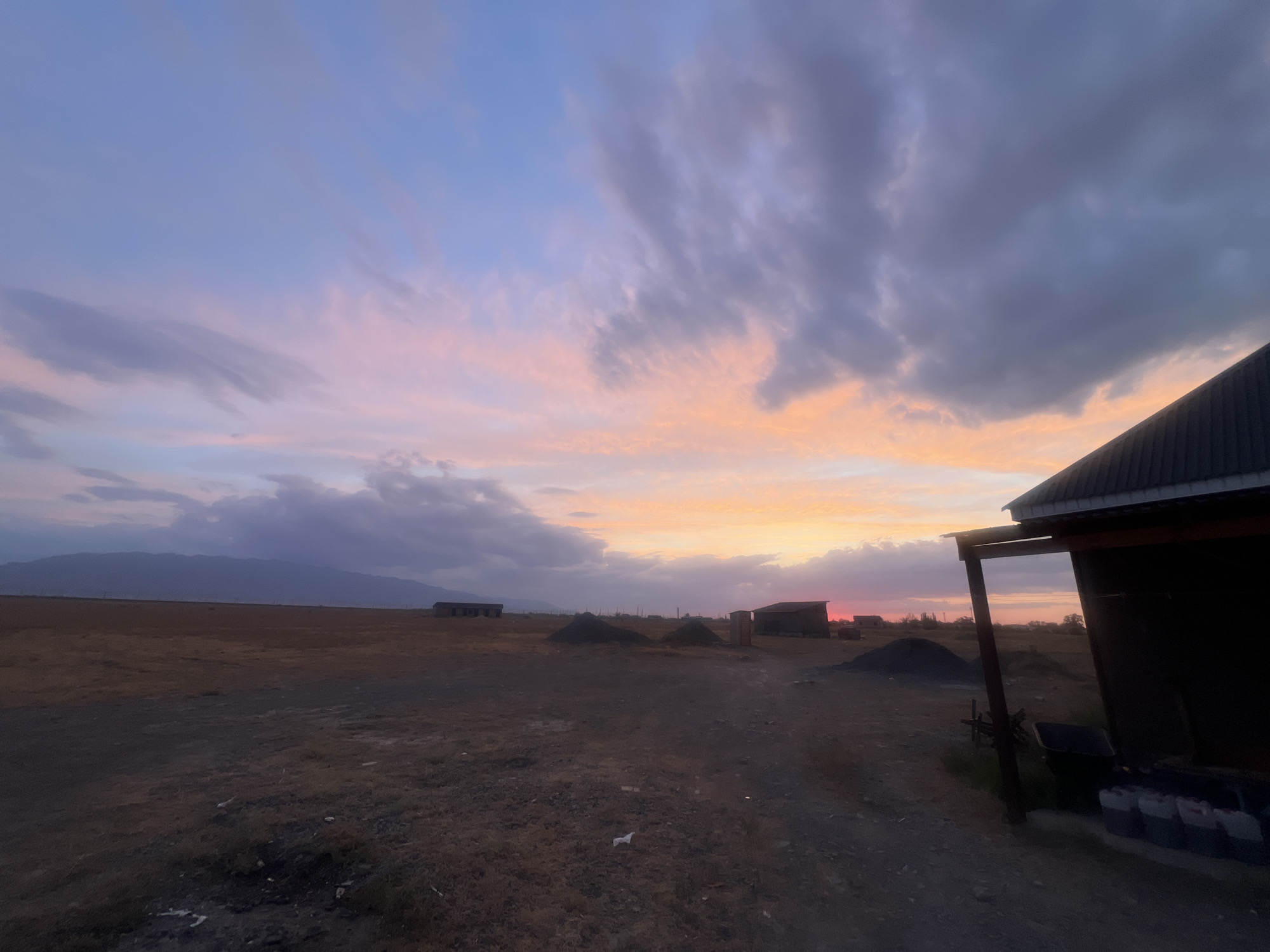 This image depicts a wide, open, and somewhat desolate landscape under a vibrant sunset sky. The foreground is dominated by dry, patchy ground with scattered debris and some small mounds of dark material. To the right, a portion of a dark, nondescript building with a corrugated metal roof is visible, with some stacked containers in front. In the mid-ground, several low-lying structures, possibly sheds or storage units, are scattered across the flat terrain, along with more conical piles of dark material. Further in the distance, a range of hazy mountains forms the horizon. The sky is a dramatic display of colors, with soft pinks and oranges blending into blues and grays as clouds drift across. The overall impression is one of a rural or industrial outskirts area at dusk. The location is identified as Ақши, Kazakhstan. There are no people or animals visible in the scene.