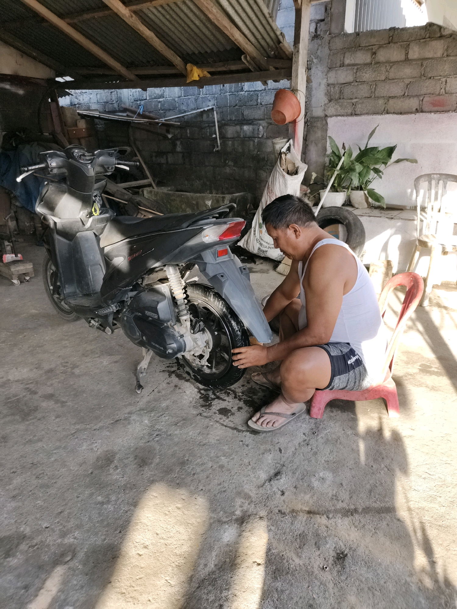 In Tombatu, Indonesia, a man is seen kneeling beside a motorcycle, which is parked on a concrete floor under a corrugated tin roof. The man, wearing a white tank top and patterned shorts, is interacting with the rear wheel of the motorcycle. The environment appears to be an outdoor covered area, possibly a workshop or garage, with brick walls and exposed wooden beams supporting the roof. Various objects are scattered around the scene, including plants in pots, a discarded tire, and a bag. The lighting suggests it is daytime, with sunlight casting shadows on the ground.