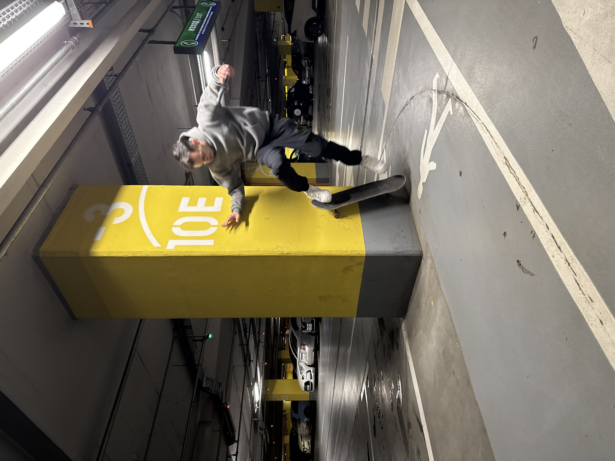 This image captures a dynamic scene of a male skateboarder performing a trick or maneuver in an underground parking garage in Sofia, Bulgaria. The photo is oriented vertically, showing the skateboarder mid-action next to a prominent yellow and gray concrete pillar. He is dressed in a gray hoodie and dark pants, with his right hand extended towards the ground or pillar and his left foot on the skateboard. His expression indicates focus and engagement in the athletic activity.
The setting is a typical underground parking facility, characterized by concrete floors marked with white lines and directional arrows, and illuminated by overhead fluorescent lighting. In the background, blurred parked cars are visible. A notable detail is a green overhead sign displaying "ИЗХОД / EXIT" in both Cyrillic and English, consistent with the specified location. The large yellow pillar, acting as a central element, features partial white numbering, including a "3" and "10E," likely denoting a parking level or zone.