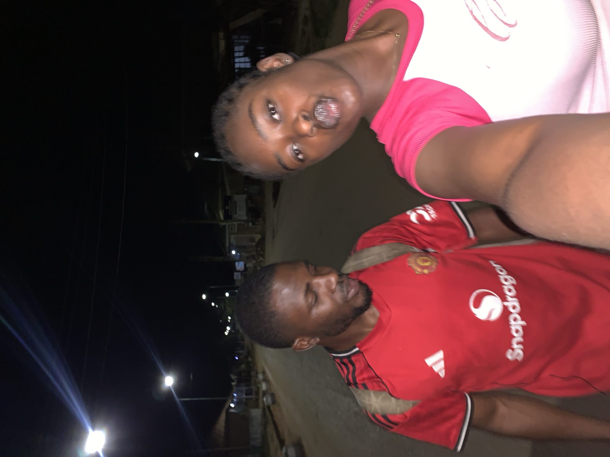 The image depicts two people at night in what appears to be a street scene. A woman with short hair is wearing a white shirt with pink sleeves. She is positioned at the top of the frame, facing the camera. A man with short hair and a beard is wearing a red Manchester United soccer jersey with "Snapdragon" written on the front. He is located below the woman. In the background, streetlights illuminate buildings and the area surrounding the New Stadium in Nigeria. The overall lighting is dark with artificial light sources.
