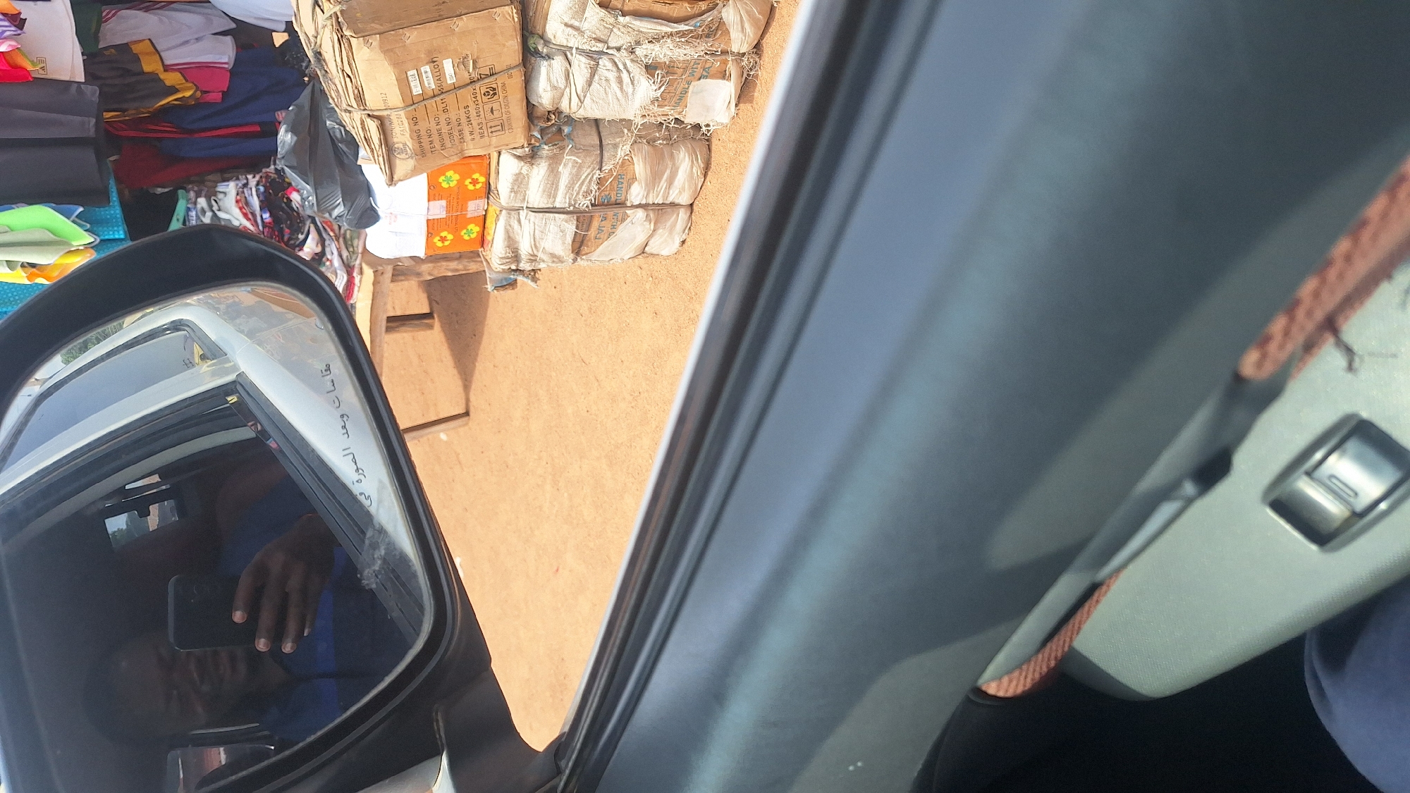 A man is reflected in a car's side mirror, holding a smartphone and taking a selfie. He is wearing a blue collared shirt. The car's interior, including a window control, is visible in the foreground. Outside the car, a market stall with various goods, including stacked boxes and bags, is visible. The ground is covered in reddish-brown soil, typical of Jalingo, Nigeria.