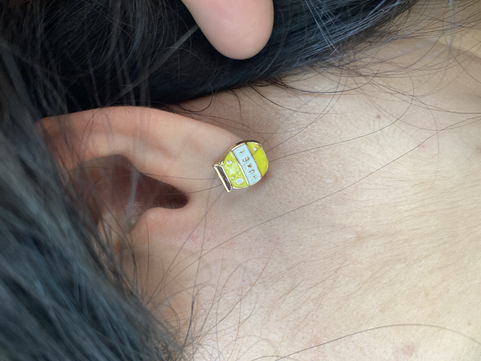 A close-up shot shows a person's ear adorned with a small, honey jar-shaped earring. The earring has a yellow and white color scheme, with the word "HONEY" written on it. The person has black hair that partially obscures the ear and some of the surrounding skin. The skin visible around the ear is fair and has some visible pores and fine hairs. The image does not provide any information to identify that the image was taken in Puebla, Mexico.