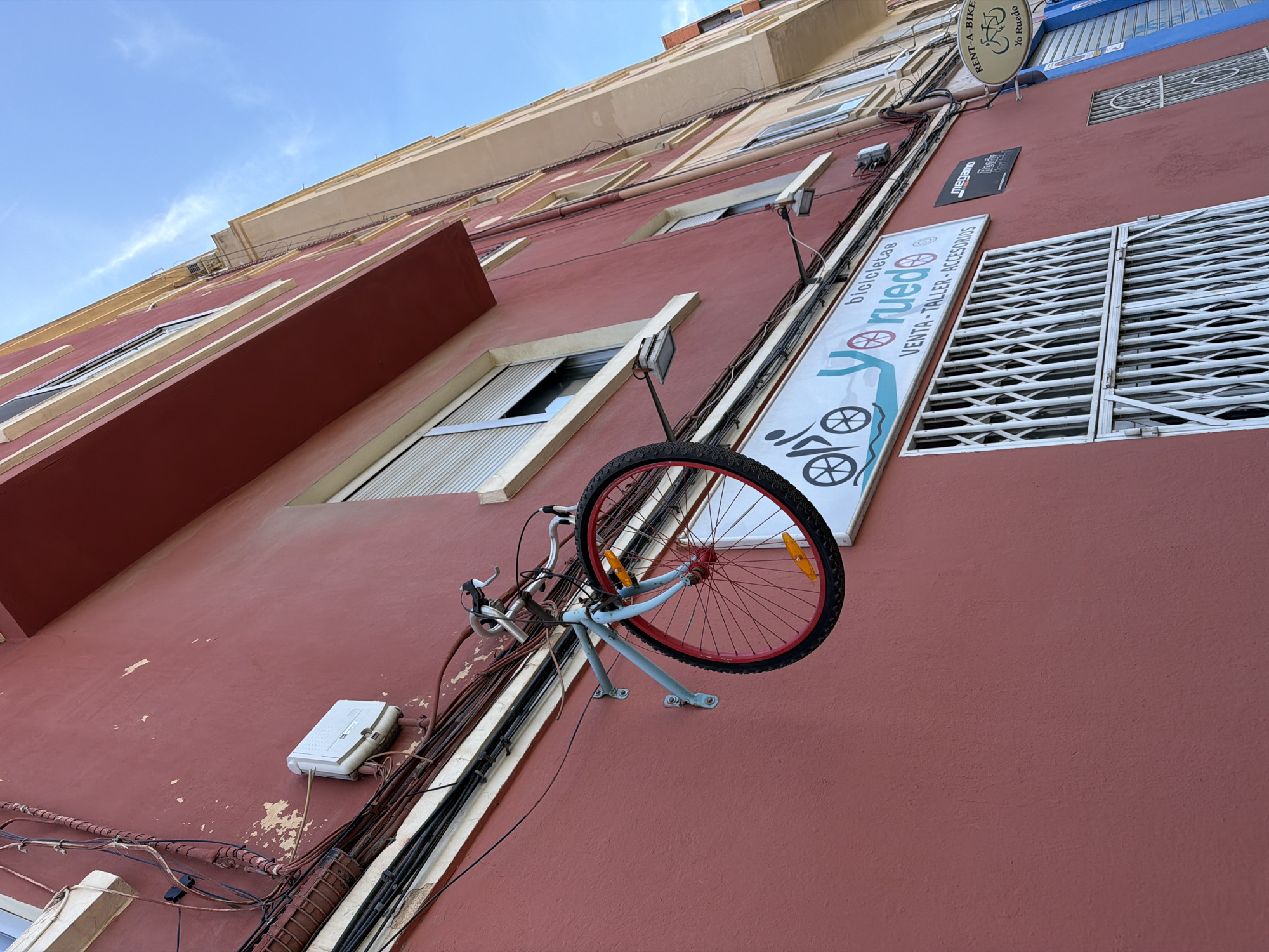 A low-angle shot reveals a red building facade in València, Spain, adorned with advertisements related to bicycles. The most prominent sign reads "bicicletas Yo Ruedo VENTA-TALLER-ACCESORIOS," indicating a bicycle shop. An incomplete bicycle, specifically its front half with a red-rimmed wheel, is mounted on the wall as part of the shop's display. The wall is also crisscrossed with electrical cables and conduits, and there is an electrical box attached to the wall. The sky is visible at the top of the frame, a light blue color with a few wispy clouds. Another sign reads "RENT-A-BIKE Yo Ruedo" with a bicycle image on the side. A closed, silver-colored metal grating covering a window is visible.