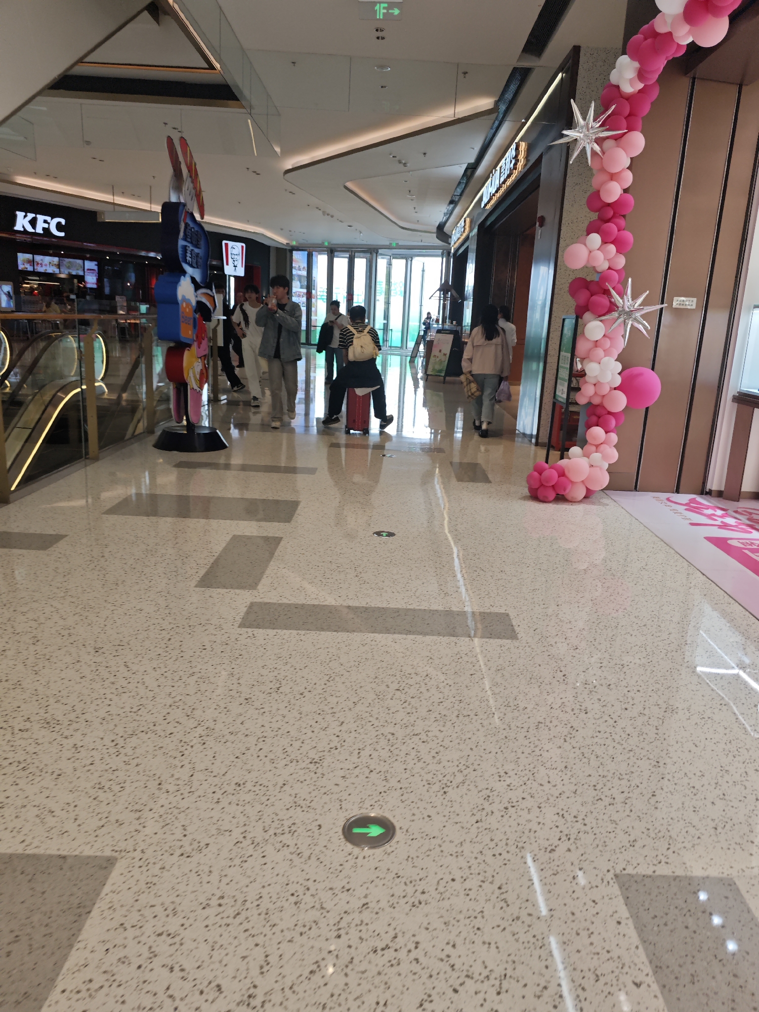 The image shows an interior scene in a shopping mall in the Liangjiang New Area of Chongqing, China. The floor is a speckled gray and white terrazzo with green directional arrow stickers affixed to it. In the background, a KFC restaurant is visible to the left. An escalator is partially visible on the left side of the image. Several people are seen walking further into the mall toward a bright, glass-enclosed entrance in the background. To the right, there is a store decorated with a pink and white balloon arch, adorned with silver starburst decorations. A tall, multi-colored decorative figure stands near the escalator.