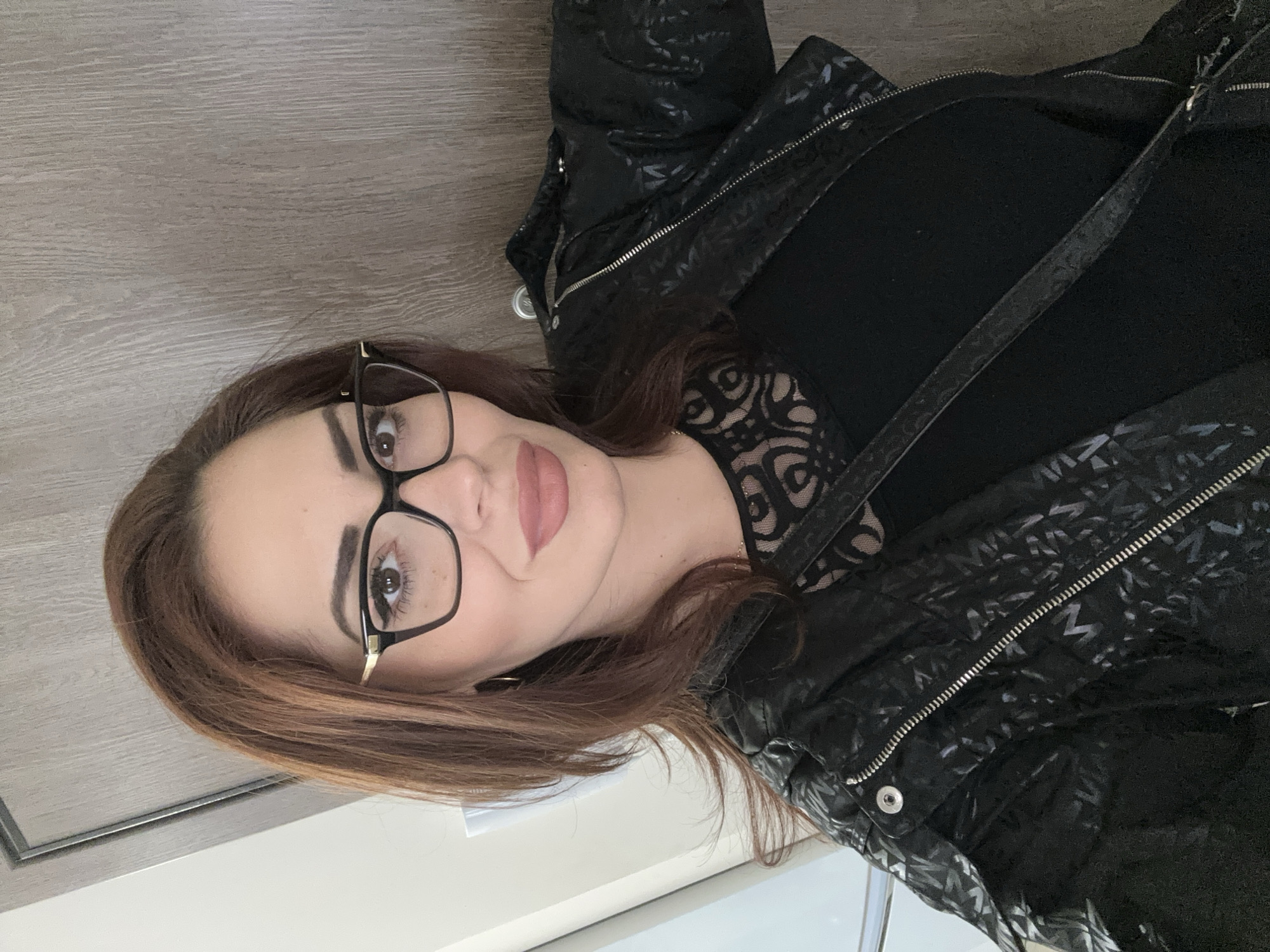 A woman with shoulder-length brown hair, wearing glasses, a black top with lace detailing, and a patterned black jacket poses for a photograph. The background consists of a light brown wood-like surface and white walls. The image does not contain enough information to determine the location in Sofia, Bulgaria.