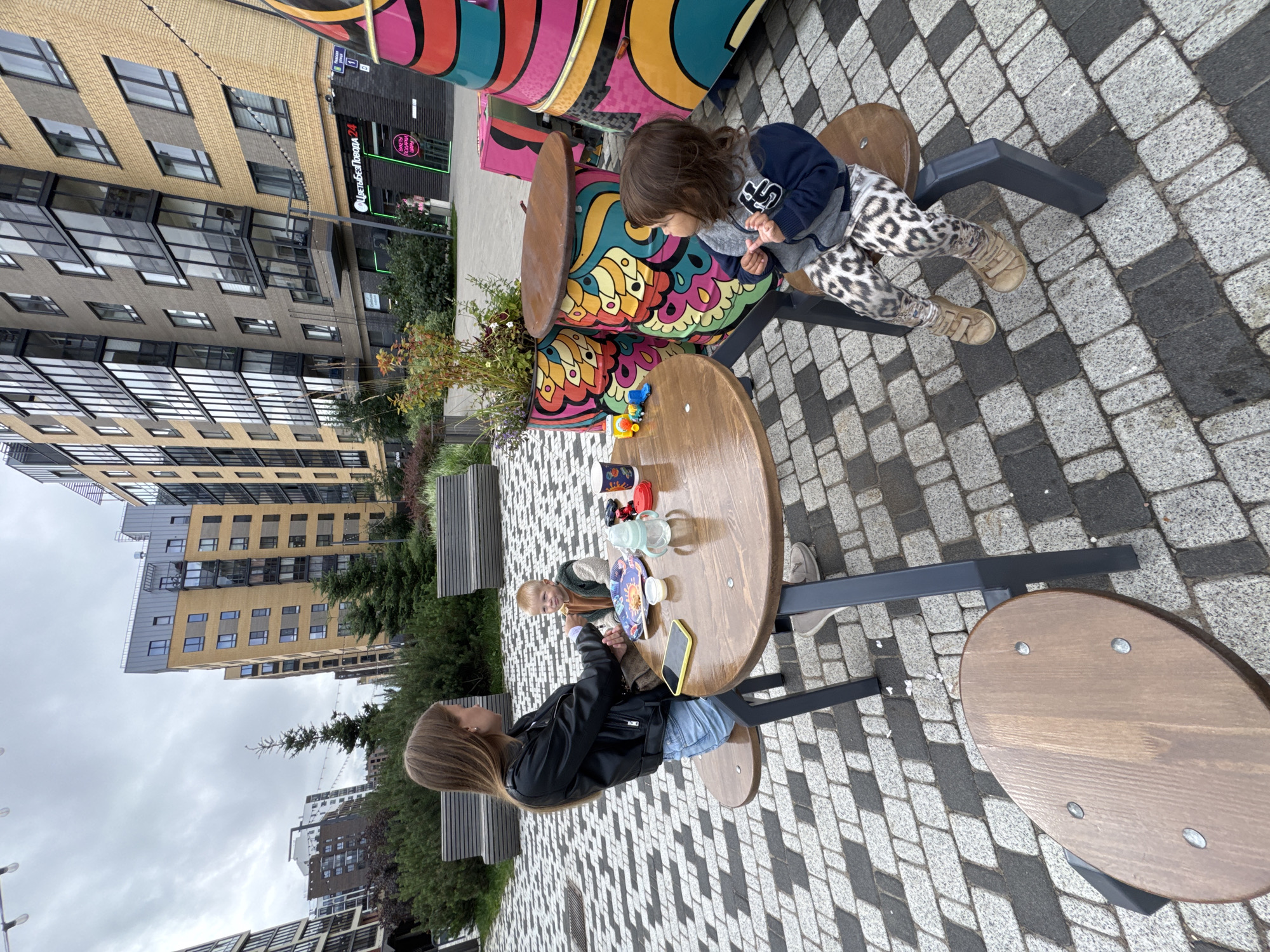 The image depicts a casual outdoor scene with three people and colorful decorative elements in a modern urban setting, likely in Novoselye, Russia. In the foreground, a round wooden table is set with plates, cups, and a baby bottle. A young woman with long brown hair, wearing a black leather jacket and denim shorts, is seated on a bench, feeding a young boy with blonde hair, who is wearing a green and brown jacket. Across the table, a young child with dark hair, dressed in a grey and navy blue jacket and leopard-print leggings, is seated and looking to the side.
The table and benches are situated on a patterned cobblestone patio. Behind the table, large, brightly colored cylindrical planters with psychedelic patterns are visible. In the background, modern apartment buildings with yellow and grey facades rise under an overcast sky. A street sign with Russian text and a number "1" is visible on one of the buildings, near a storefront. The overall environment suggests a developed residential area with communal outdoor spaces.