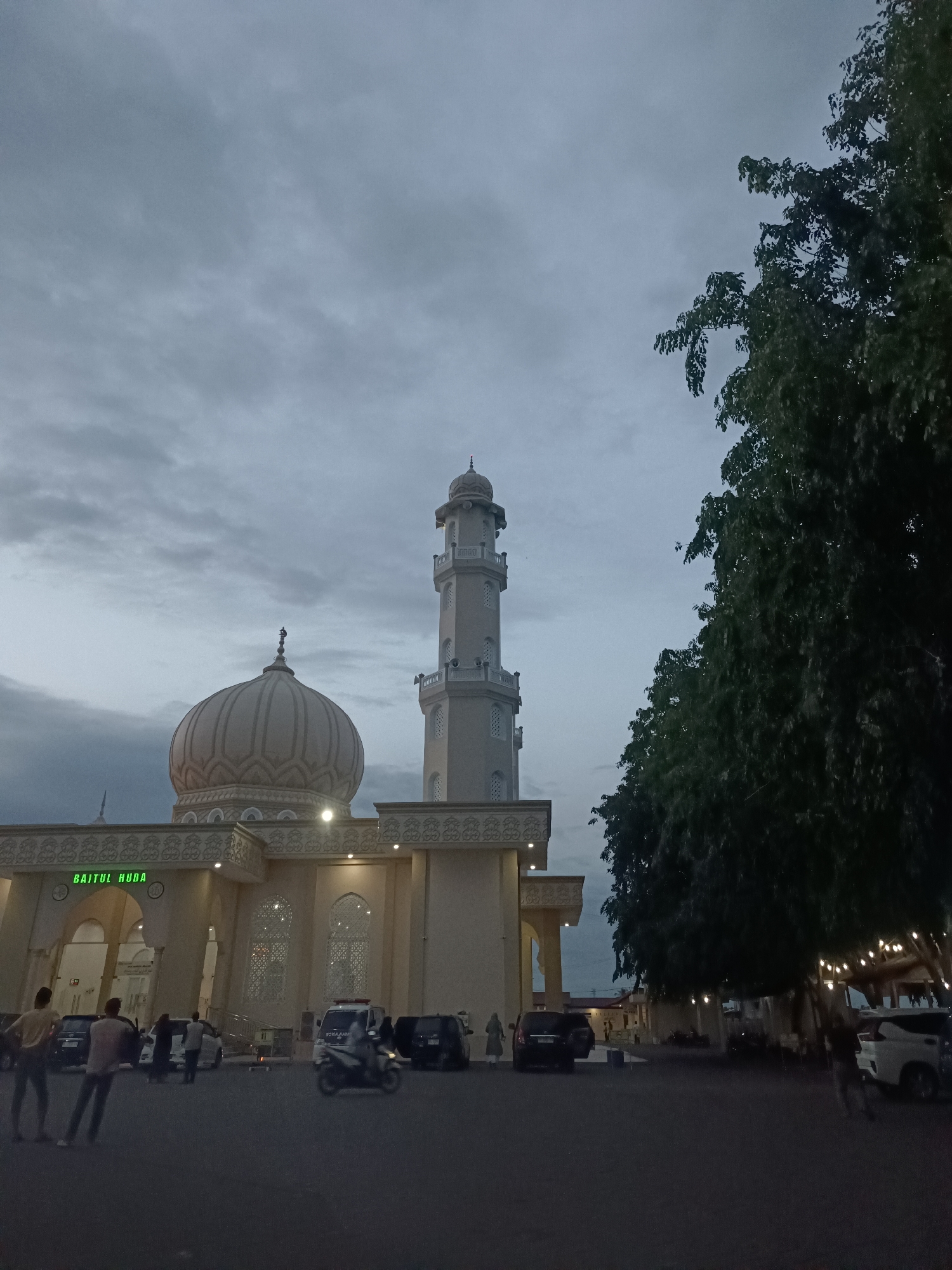 The image depicts the Baitul Huda Mosque in the city of Mee Tanjong Usi, Indonesia, during twilight. The mosque is a light-colored structure with a prominent dome and a tall minaret. The sky above is overcast with gray clouds. In front of the mosque, there is a paved area with several parked cars and a motorcycle. A few individuals are visible, some standing and walking. To the right of the mosque, a large tree with dark green foliage is prominent, and string lights are visible hanging from branches. The overall scene is dimly lit, characteristic of early evening or late afternoon before complete darkness.