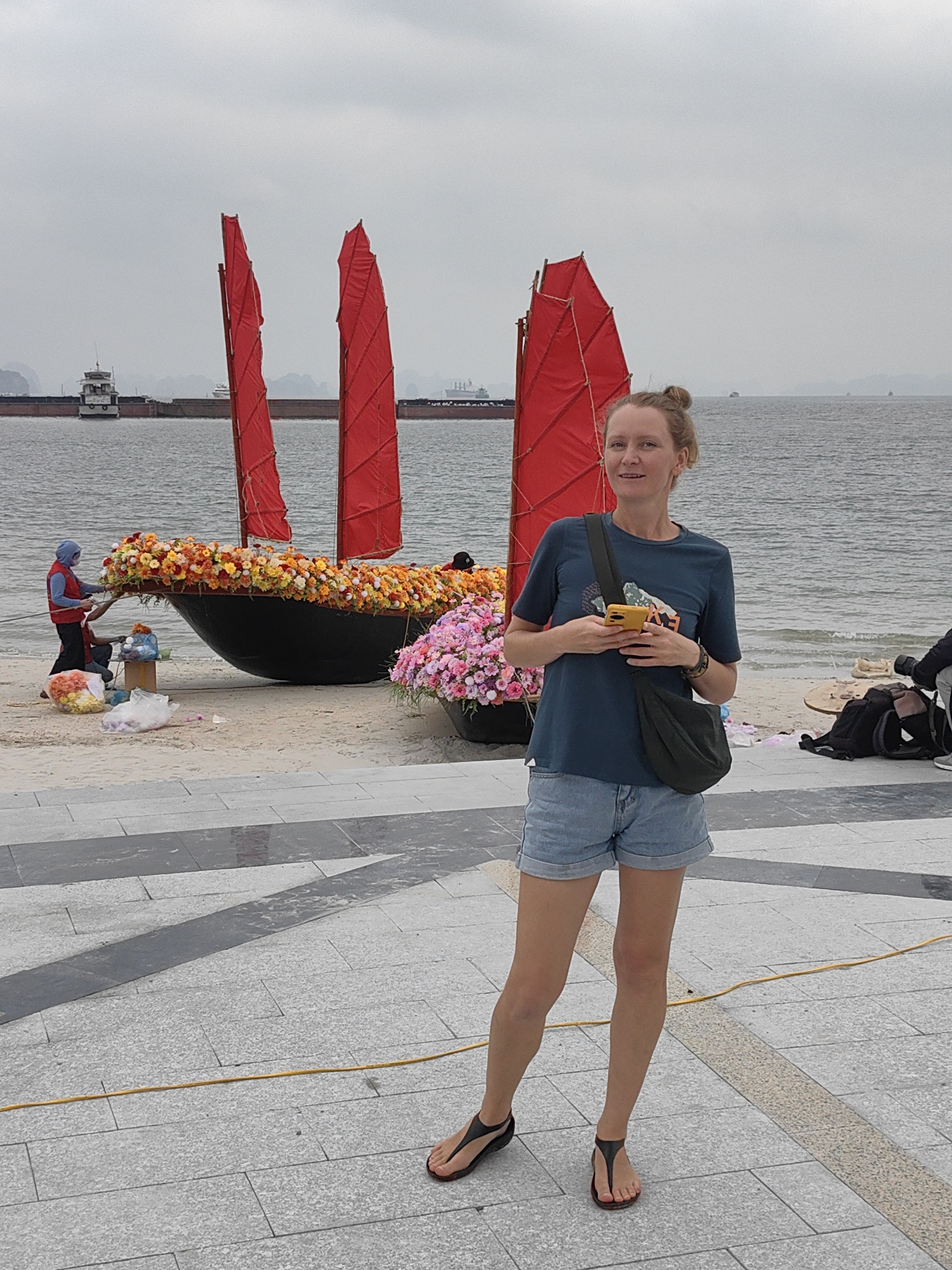 A woman stands on a paved walkway in Bai Chay Ward, Vietnam. She wears a blue t-shirt, jean shorts, and sandals, and holds a yellow phone. Behind her is a beach with a black boat decorated with flowers and three red sails. To the left of the boat, two individuals are working with flowers. A large ship is visible on the water in the background. The sky is overcast. A yellow cable runs along the pavement in the foreground.