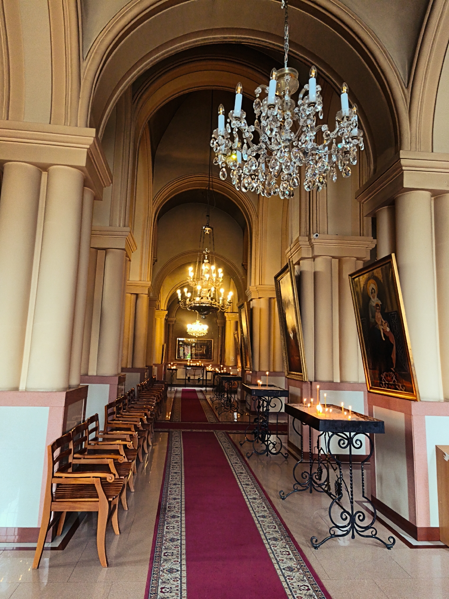 This is an interior view of a church, possibly a cathedral, in Vladikavkaz, Russia. The scene is captured from the perspective of someone standing near the entrance and looking down a long aisle. The architecture features tall, rounded arches supported by thick, fluted columns painted in a light, peachy color. Chandeliers made of crystal hang from the ceiling.
The aisle is lined with wooden chairs or pews on the left. On the right, a series of ornate black metal tables hold numerous lit candles, casting a warm glow. Framed religious paintings hang on the walls, leaning against the columns. A plush red carpet with an intricate patterned border runs down the center of the aisle. The lighting is warm and atmospheric, suggesting either natural light filtering in or the effect of the candles and chandeliers. No people are visible in the frame.
