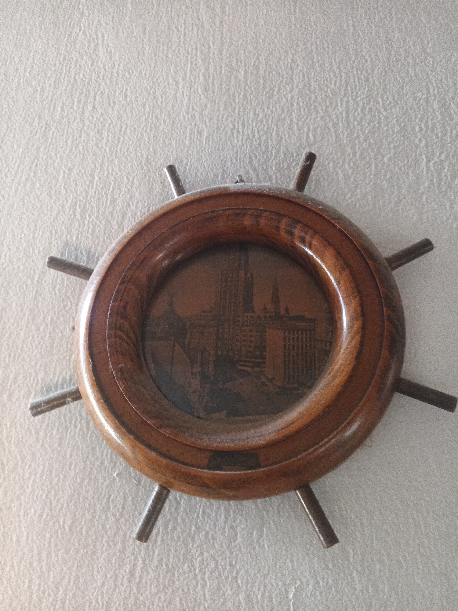 A wooden decorative object shaped like a ship's wheel hangs on a textured off-white wall. The wheel's central area contains a circular image of a cityscape, including a tall building with a spire. The cityscape is a view of Caracas, Venezuela, specifically the Parque Central Complex.