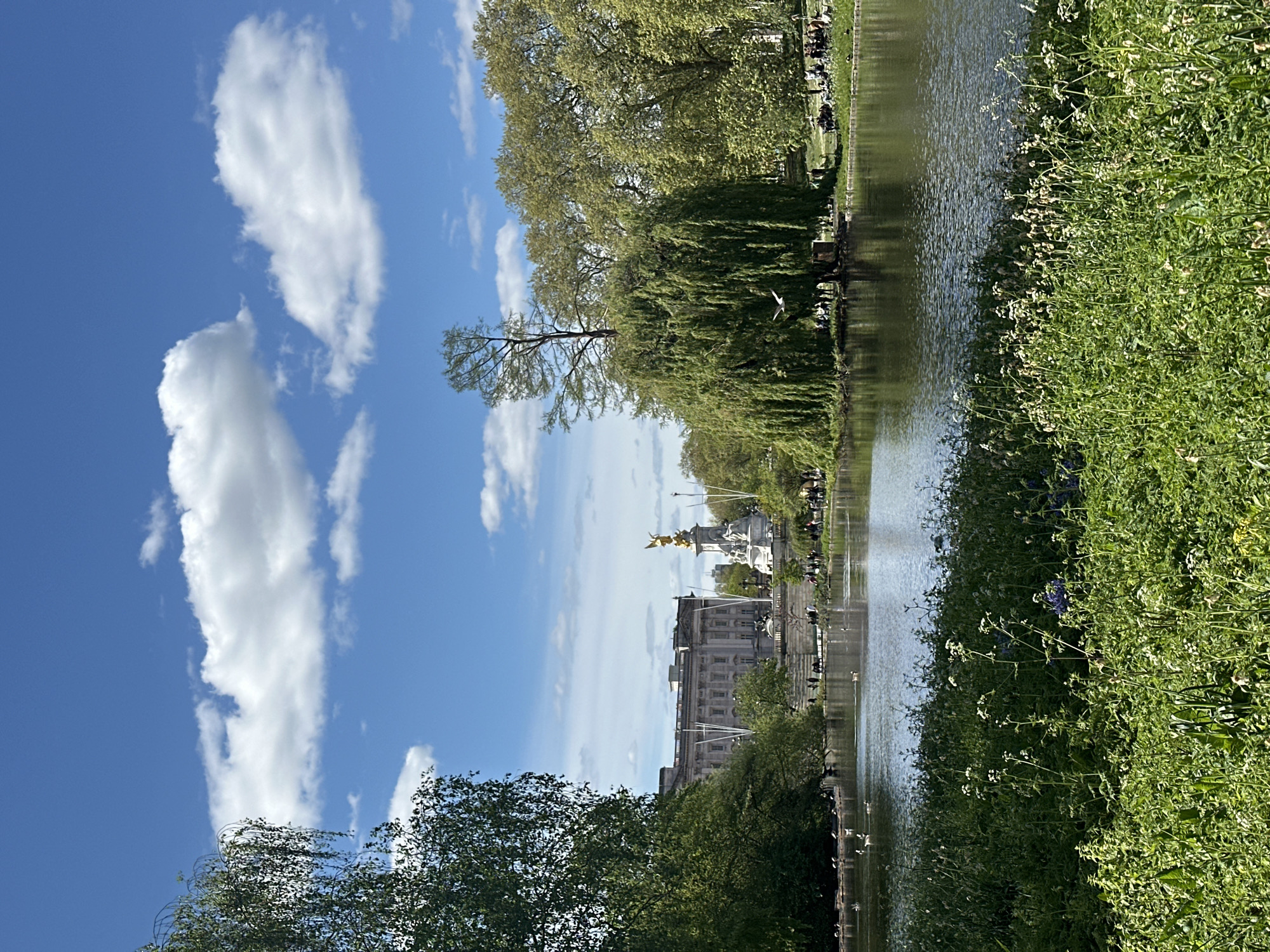 This image shows a view of Buckingham Palace in London, United Kingdom, across a body of water, likely St. James's Park Lake. The Palace, a large building with a statue on top, is partially obscured by trees. The sky is blue with large, scattered white clouds. The lake is flanked by lush green vegetation on one side. There are people along the bank of the lake in the distance.