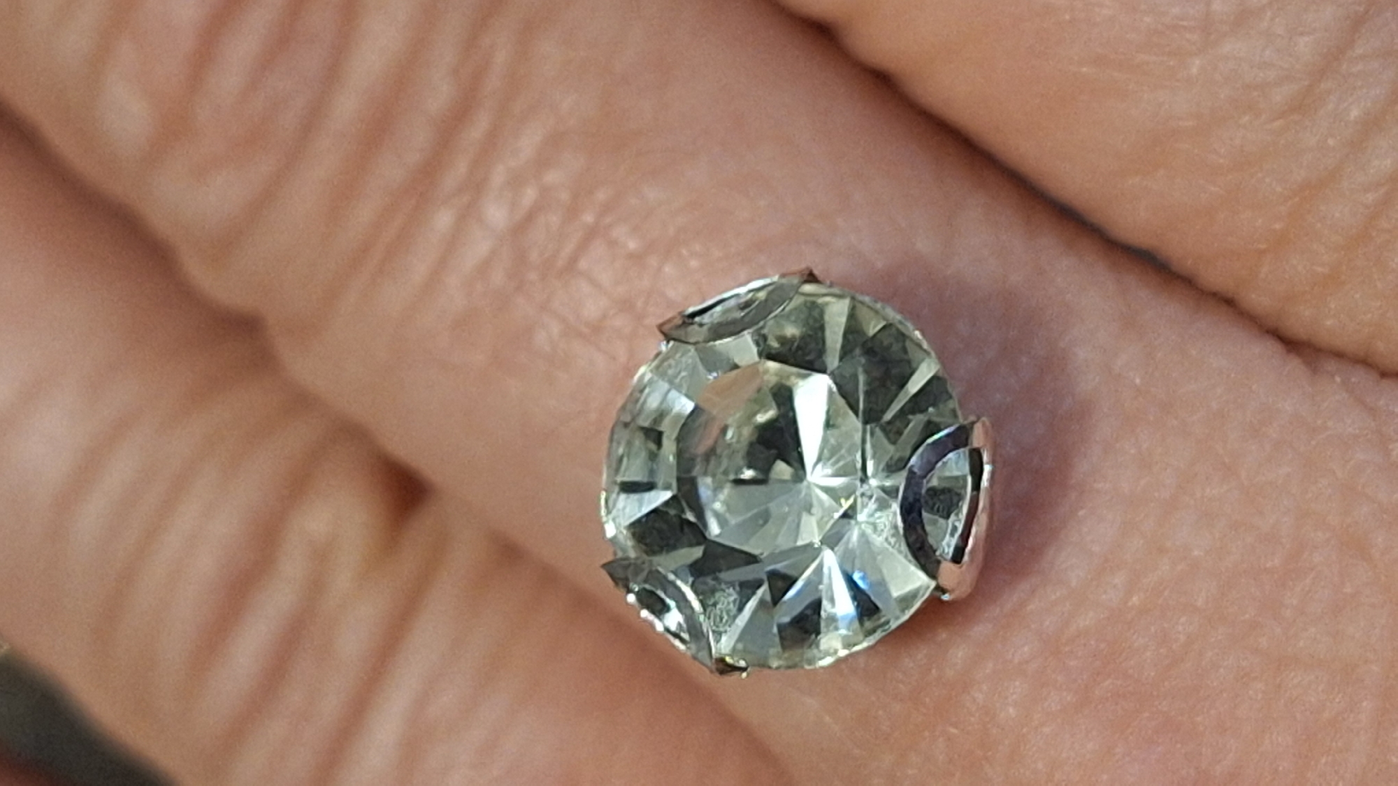 A close-up, high-angle shot captures a faceted, clear or very light-colored gemstone, possibly a diamond or crystal, held against the textured skin of a hand. The round gemstone is prominently displayed in the center of the frame, secured by a four-prong metal setting, which appears to be silver or white gold. Light reflects brightly off the numerous facets of the stone, showcasing its intricate cut. The surrounding skin of the hand, likely the palm or inner fingers due to the visible creases and lines, serves as the background, filling the entire frame with its warm, light-to-medium tone. There are no other discernible objects, activities, or text visible in the image. The setting appears to be indoors or in a well-lit environment.