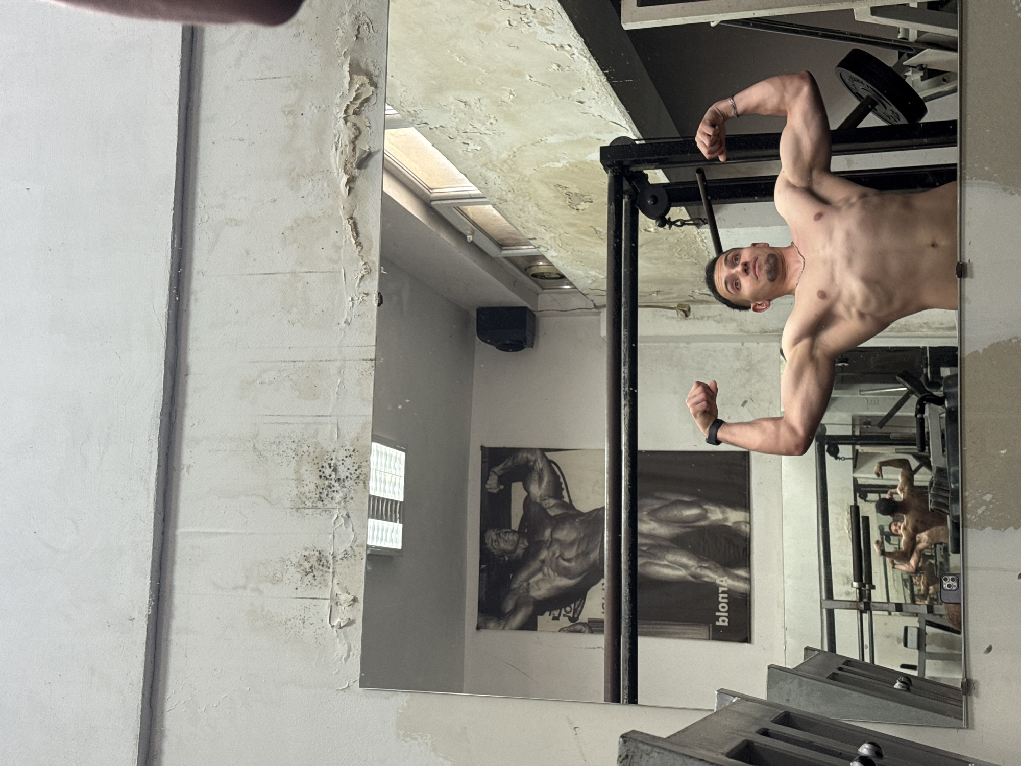A bare-chested man flexes his arms in front of a mirror. He wears a bracelet on his left wrist and a necklace. The mirror reflects a weightlifting machine and a poster featuring an image of a bodybuilder. The wall behind the mirror is deteriorating, with peeling paint and mold. The location may be in a gym or fitness studio in Shumen, Bulgaria.