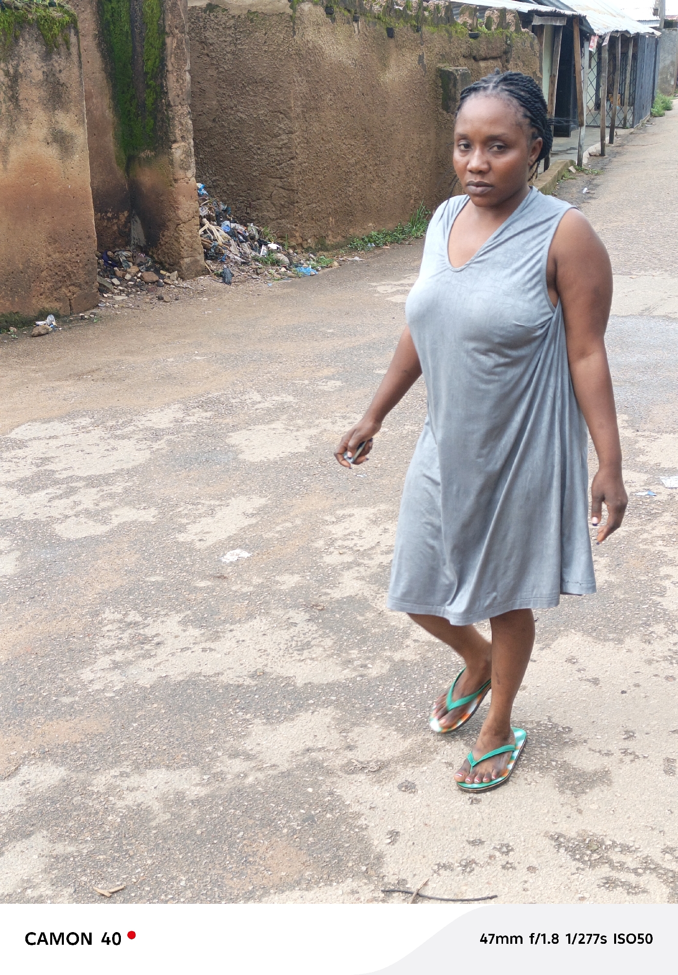 A woman with braided hair, wearing a gray sleeveless dress and patterned flip-flops, walks on a paved street. She is looking towards the camera. The background features a brown stucco wall with moss growing in some areas, and a pile of garbage at the base of the wall. Further in the background, there are buildings with corrugated metal roofs. The ground is a worn asphalt surface with patches of lighter gray concrete. The image is a full-body shot of the woman, with her from the chest down being more in focus. The scene appears to be in an outdoor, urban environment.