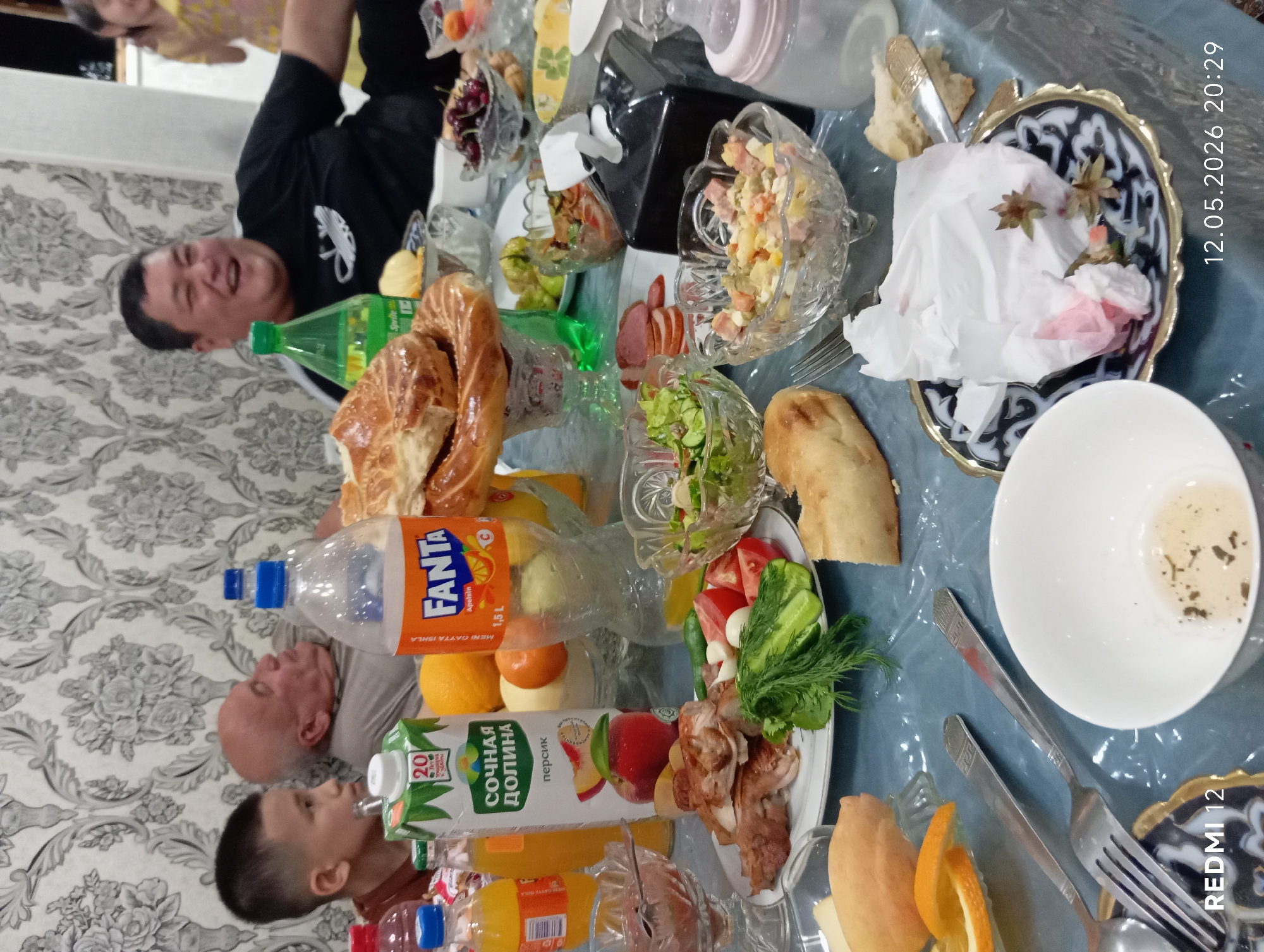 The image depicts a family meal in what appears to be a private residence. Several individuals are visible around a table laden with food and drinks. The table is covered with a light blue tablecloth and features a variety of dishes including salads, bread, pastries, sliced meats, fresh fruits such as oranges, and beverages like Fanta and peach juice. There is also a baby bottle present. The individuals visible include a smiling man in a black shirt, a boy drinking juice, and another man with a bald head. The setting appears to be indoors, with a patterned wallpaper visible in the background. Based on the OCR text, the scene may be located in the city of Bekabad, Uzbekistan, as the Fanta bottle has text "MENI GAYTA ISHLA," likely indicating it is being produced locally in Uzbekistan. The date 12.05.2026 20:29 appears on the right of the image.