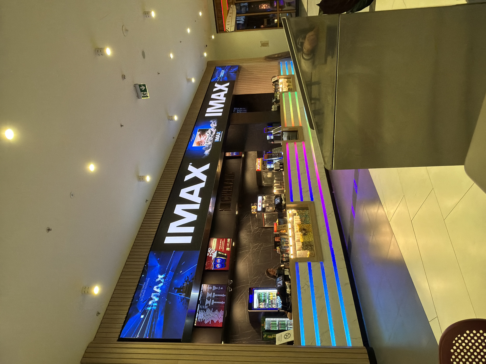 The image depicts the IMAX concession stand in a shopping mall in Plovdiv, Bulgaria. The prominent "IMAX" logo spans a series of screens displaying movies. Below the screens, various displays show movie schedules and food/drink items for sale. The concession stand itself features rows of snacks, popcorn, drinks, and point-of-sale systems manned by employees. Colorful vertical LED strips decorate the stand. The setting suggests an indoor mall environment, with a chair visible in the lower right corner.