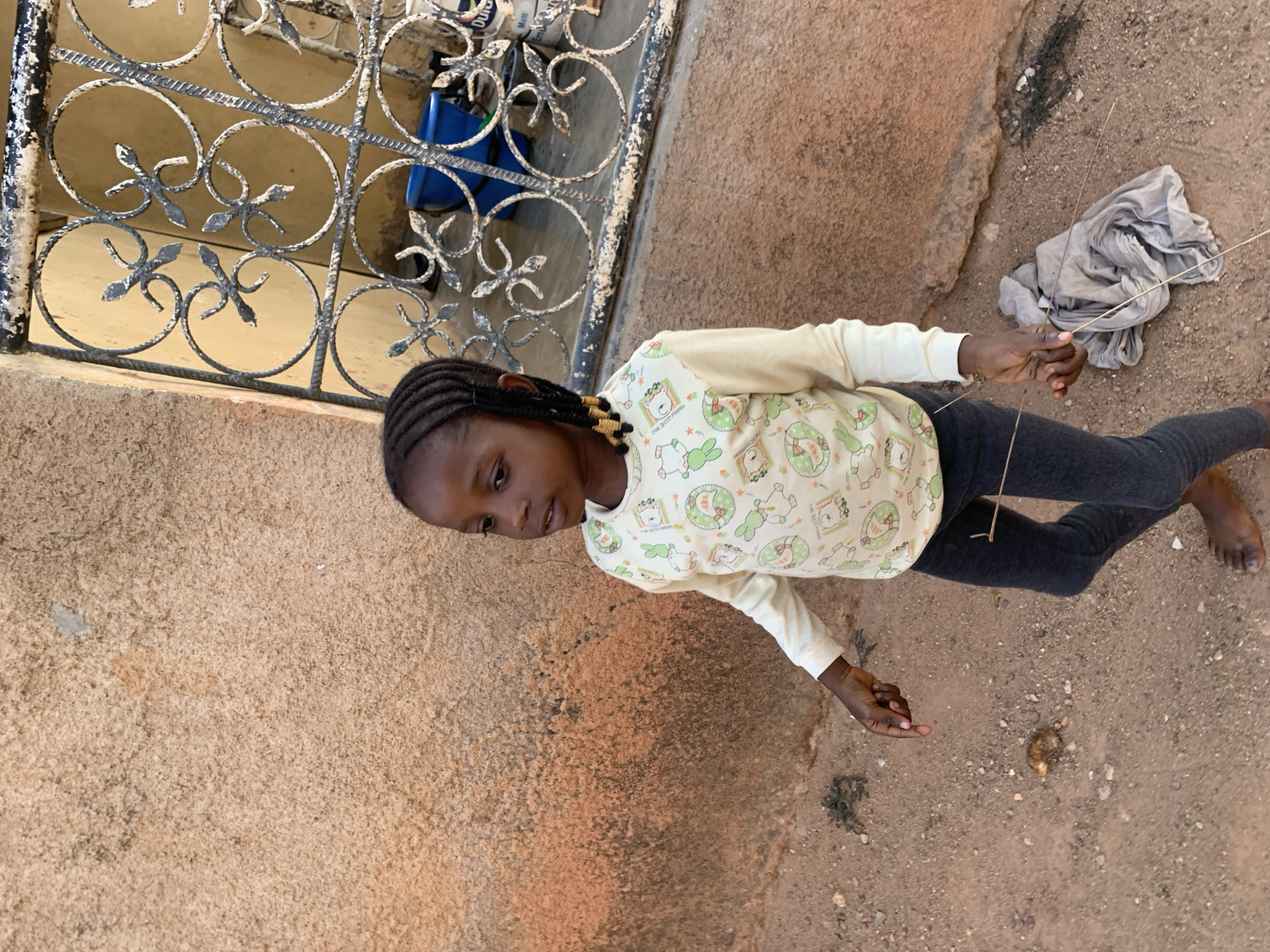 A young Black girl with braided hair stands barefoot on a dirt surface. She wears a patterned long-sleeve shirt and grey leggings. To her left is a weathered, ornate metal gate. Next to her, on the ground, is a crumpled grey cloth and two sticks. The surface she stands on is a mix of dirt and sand. The background includes a textured, light brown wall or building exterior. The location appears to be outside. There is no definitive visual information in the image that confirms it is Jos, Nigeria.