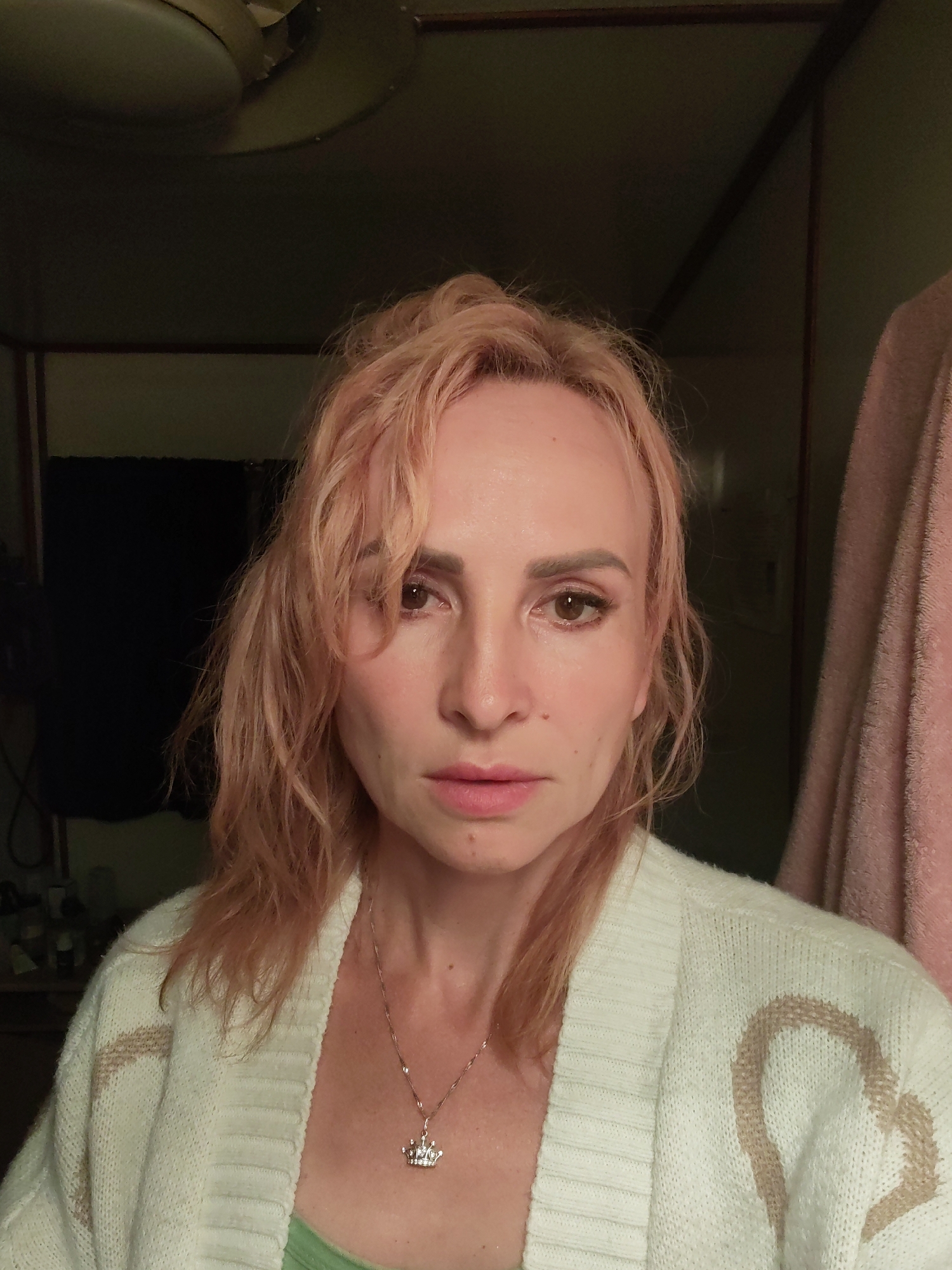 The image shows a close-up, front-facing, slightly overhead view of a person with blonde-pink hair. The person is wearing a cream-colored knitted cardigan with a tan heart pattern, and a light green undershirt. A delicate silver necklace with a crown-shaped pendant is visible on their neck. The person's gaze is directed towards the camera, and their expression is neutral. The background is dimly lit and appears to be an indoor environment, possibly a bedroom or dressing area, with hints of dark fabric, a ceiling fixture, and a pink textile hanging to the right. There is no discernible location within the city of Shalsky, Russia, indicated in the image.