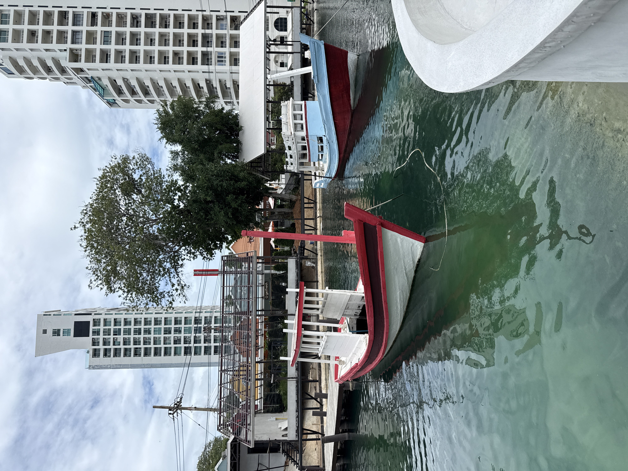 The image shows a canal in Pattaya City, Thailand, with two small boats docked. The boat in the foreground is red and white, with a white wooden structure on top. The boat in the background is blue and red, also with a white wooden structure. Both boats are floating on green water. In the background, there are tall, modern buildings, some of which have curved balconies. There are also trees and some smaller structures, possibly shops or restaurants, along the canal. The sky is overcast with clouds.