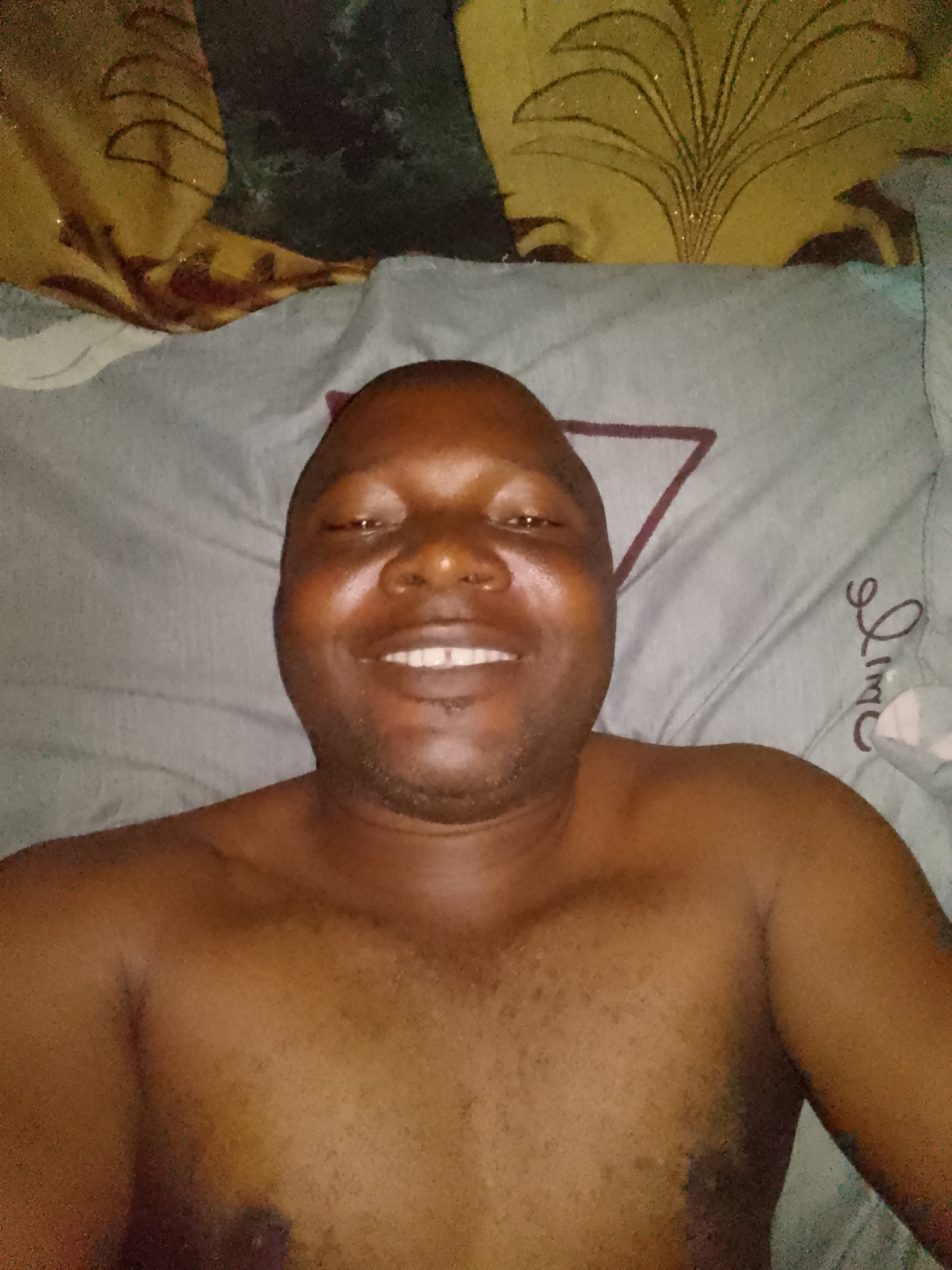 A bald man with dark skin is captured in a close-up, chest-up shot, likely a selfie. He is lying down, shirtless, with his shoulders and upper chest visible. He has a broad, toothy smile, indicating a happy and relaxed mood, and is looking directly at the camera. His face shows some shine, possibly from skin oils or lighting.
He is resting on what appears to be a bed. Directly beneath his head and extending to his right is a light grey pillow with a dark reddish-brown triangular design. To the right of his head, on the same pillow, cursive text is visible, which appears to spell "Lime" or a similar word. Above his head is another piece of bedding, possibly a pillow or blanket, that is yellowish-gold with a stylized, glittery floral or leaf-like pattern. The background is slightly out of focus, emphasizing the man. The lighting suggests an indoor setting, possibly with artificial light, given the slight graininess of the image.