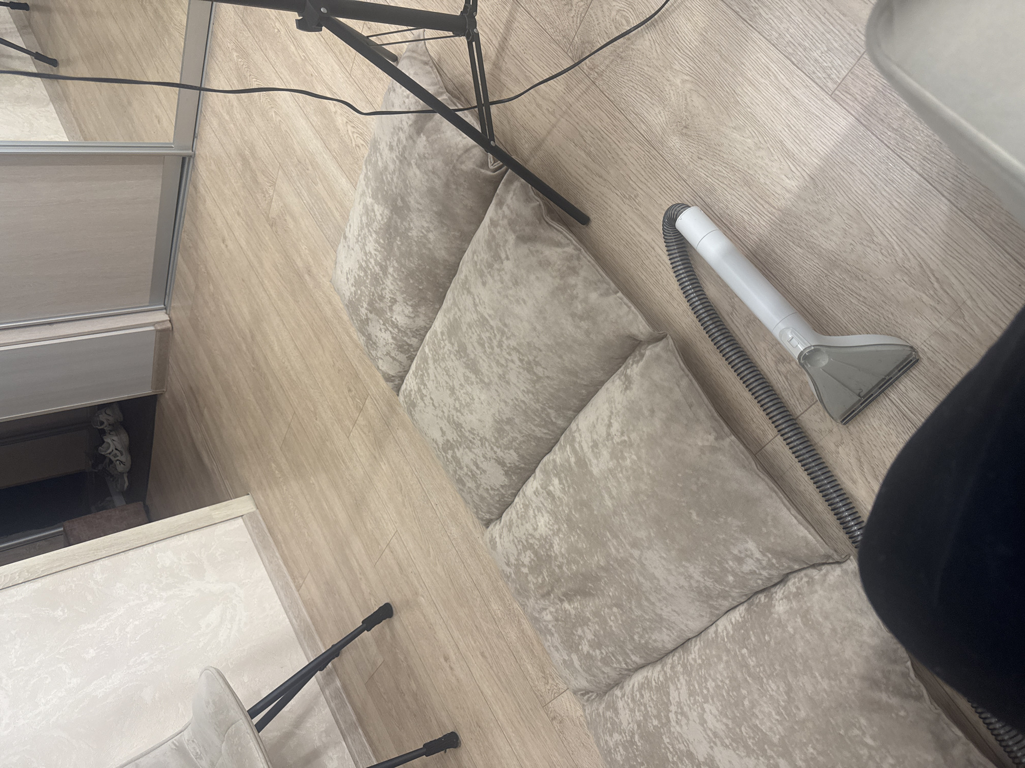 The image depicts an indoor scene, likely a living space, with light-colored wooden flooring. In the foreground, a person's legs are visible, clad in dark pants. Lying on the floor are several large, rectangular floor cushions with a textured, light beige fabric. A vacuum cleaner hose with a clear plastic attachment is positioned near the cushions, suggesting a cleaning activity is underway or has recently occurred. In the background, a chair with black legs and a light-colored cushioned seat is partially visible. Further back, a doorway leads to another room where a pair of white athletic shoes are placed on the floor. A wardrobe with mirrored doors is also visible in the distance. A tripod with a black cord is present on the right side of the frame. The overall impression is one of a domestic environment during a cleaning or tidying process. The image does not provide any specific information to identify the location as Saratov, Russia.