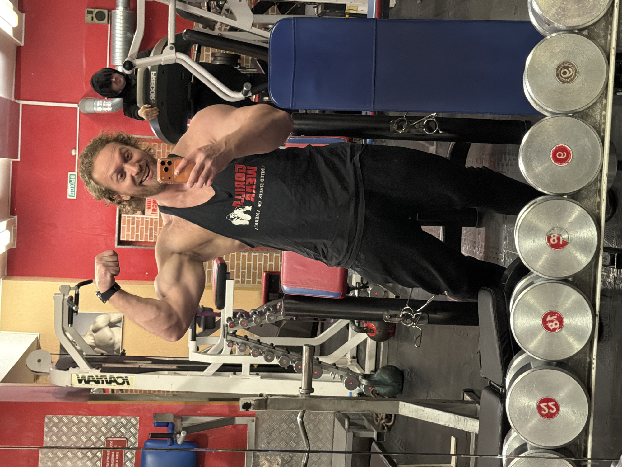 The image depicts an indoor gym environment, likely located in Saint Petersburg, Russia, based on the provided context.
In the foreground, a muscular male subject with curly blonde hair and a beard is captured in a mirror selfie. He is smiling broadly, flexing his right bicep, and holding an orange smartphone in his left hand, which is equipped with a dark-colored smartwatch. He wears a black tank top with a white "GORILLA WEAR UNITED STATES OF AMERICA" logo and black athletic pants. Below him, in the mirror's reflection, a row of silver dumbbells with red circular weight markers is visible.
In the mid-ground, a second individual is partially visible to the right of the main subject. This person is wearing a dark hooded sweatshirt, obscuring most of their face, and is positioned near a grey "PRECOR" branded treadmill.
The gym features red walls, with a section of exposed light-colored brickwork visible on the left. A white metal textured plate is affixed to the red wall. Above the main subject's head, a mirrored "ВЫХОД" (EXIT in Russian) sign is present. Various pieces of gym equipment populate the background, including a white multi-station cable machine, a dumbbell rack with smaller weights, a teal kettlebell, a black and red kettlebell, and blue and red upholstered gym benches. White ventilation ducts run along the upper portion of the red wall. The flooring appears to be dark-colored gym matting. Overhead lighting illuminates the space.