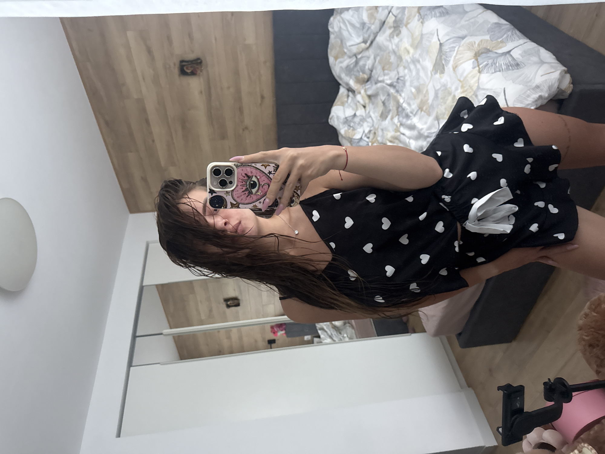 An indoor mirror selfie captures a young woman from the chest up, her reflection slightly off-center and rotated. She stands before a large mirror, holding a smartphone with both hands to take the picture. Her face is partially obscured by the phone, but her brown, wet hair, possibly just washed, cascades over her shoulders. She is wearing a black top or camisole and matching shorts or a skirt, both adorned with small white heart patterns. A white bow detail is visible on her shorts/skirt. She accessorizes with a delicate silver necklace featuring a small circular pendant and a red string bracelet on her right wrist.
The smartphone she holds has a distinctive decorative case: a light-colored (possibly pink or beige) background with a large "evil eye" design in the center, surrounded by smaller star and moon motifs. The phone's camera lenses are visible on the top left of the case.
The background of the reflection reveals a spacious room with light brown wooden flooring. A dark grey bed or sofa is visible, covered with a white and grey patterned duvet featuring leaf or floral designs, with hints of yellow or beige. A round, white light fixture is mounted on the wall to the left of the woman. To her right, a portion of a white cabinet with horizontal silver handles is reflected in the mirror. In the foreground of the reflection, a black phone stand or tripod is visible, along with parts of a pink object and a fluffy brown object, possibly a teddy bear or cushion. The overall setting suggests a bedroom or dressing area within a home. No visible text is present in the image.