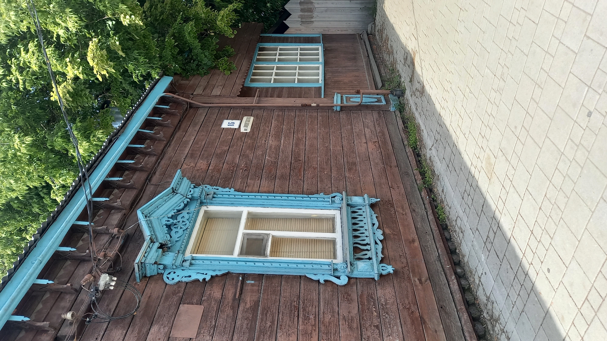 The image depicts the exterior of a weathered, brown wooden building, specifically located at 15 Dmitrovskaya Street (ул. Дмитровская) in Zvenigorod, Russia, as indicated by a white sign affixed to the wall. The building features two windows. The prominent left window has a white frame and visible internal blinds, surrounded by an elaborate, carved light-blue wooden trim (nalichniki) with decorative elements above and below. To its right, a smaller window with a blue and white multi-pane frame is set into a recessed section of the wall, with a blue-painted downspout or pipe running vertically beside it.
Above the main window, the roofline exhibits a light-blue painted fascia board with decorative carved wooden elements hanging below it, consistent with traditional Russian architectural style. Electrical wires are strung across the upper portion of the building, connected to various insulators and a white junction box mounted on the brown wooden planks. Green foliage from trees is visible above and behind the roofline, suggesting a natural environment surrounding the structure.
The ground in the foreground is a light-colored paved sidewalk composed of square interlocking pavers. Distinct shadows are cast across the sidewalk, indicating bright daylight. Sparse green vegetation grows along the base of the building's wooden wall. On the far right, a segment of a light-colored wooden picket fence is partially visible. No people are present in the scene.