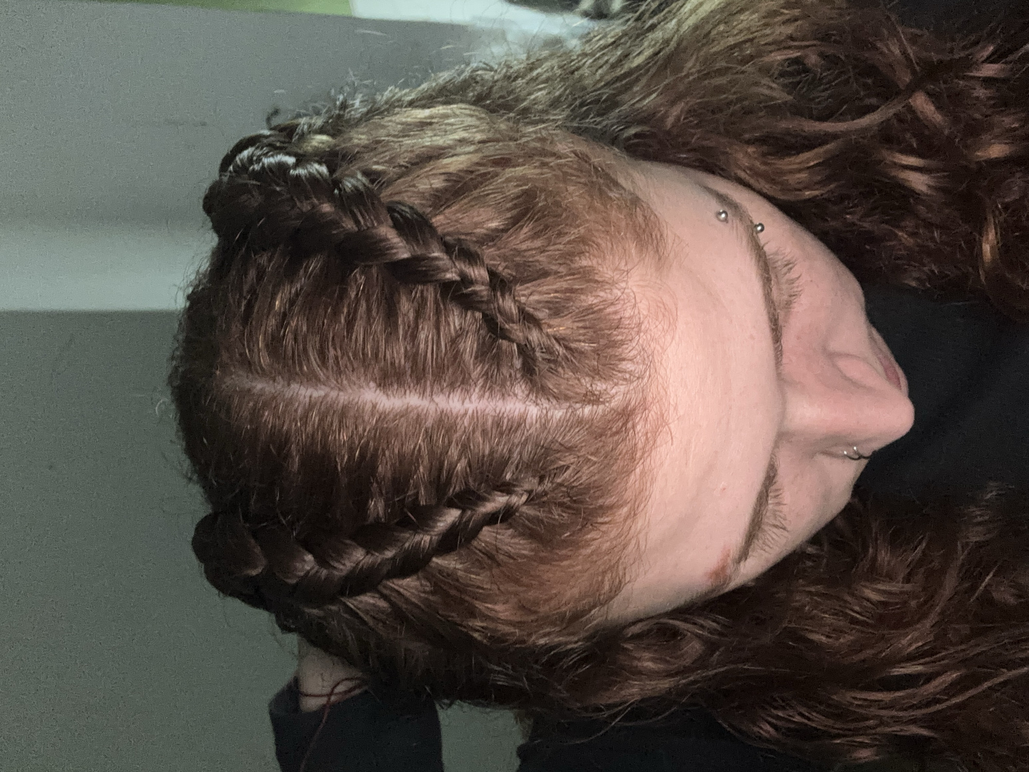 The image is a close-up, slightly downward-angled view of a person's head. The individual has light skin and reddish-brown hair styled in two braids that run along the scalp. The face is partially visible, revealing a portion of the nose and cheek. There are two piercings visible on the eyebrow. The background is a blurred mix of light and dark shades, suggesting an indoor setting. There is no information available in the image to establish a location in Buenos Aires, Argentina.