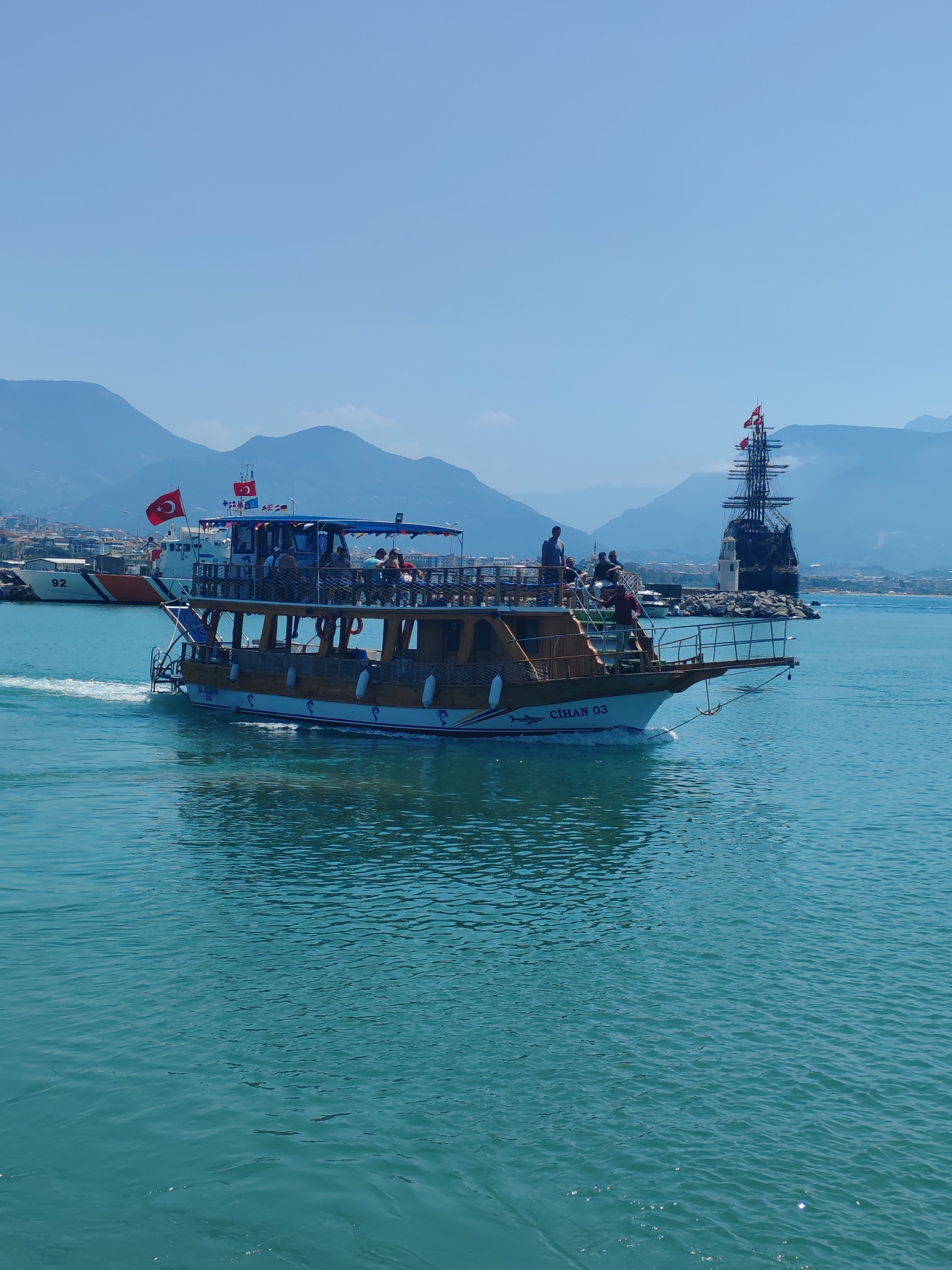 The image shows a two-story wooden tour boat, named "CIHAN 03," moving on turquoise water in Alanya, Turkey. The boat is carrying passengers on both levels. It is moving from left to right, leaving a white wake. In the background, there are mountains, the city of Alanya, a white and orange Coast Guard boat marked '92' and a black pirate ship replica standing on a breakwater. The Turkish flag is flying on the tour boat and the Coast Guard vessel, and multiple flags are flying from the pirate ship masts. The sky is clear and blue.