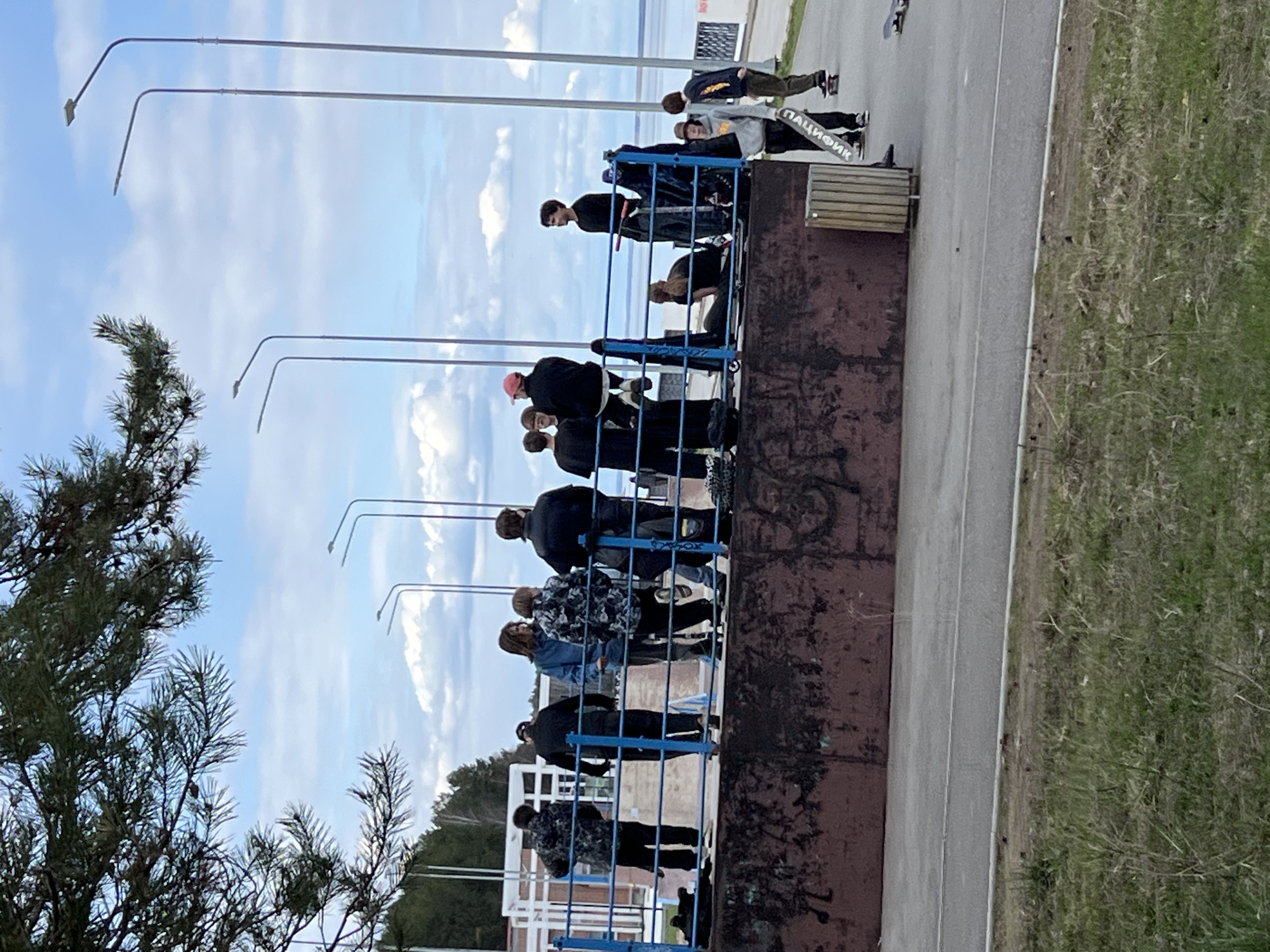 The image depicts a group of approximately 15 people standing behind a blue railing, possibly overlooking a lower area or a view. The location appears to be an outdoor area, possibly a public space or a viewing platform, indicated by the presence of streetlights, trees, and what seems to be a concrete pathway or road.
The group is composed of people of varying heights and builds. Most are dressed in casual clothing, including jackets, jeans, and sweaters.
Based on the skateboard with the inscription "ПАЦИФИК" (Pacific), the location is identified as the city of Chaikovsky, Russia.