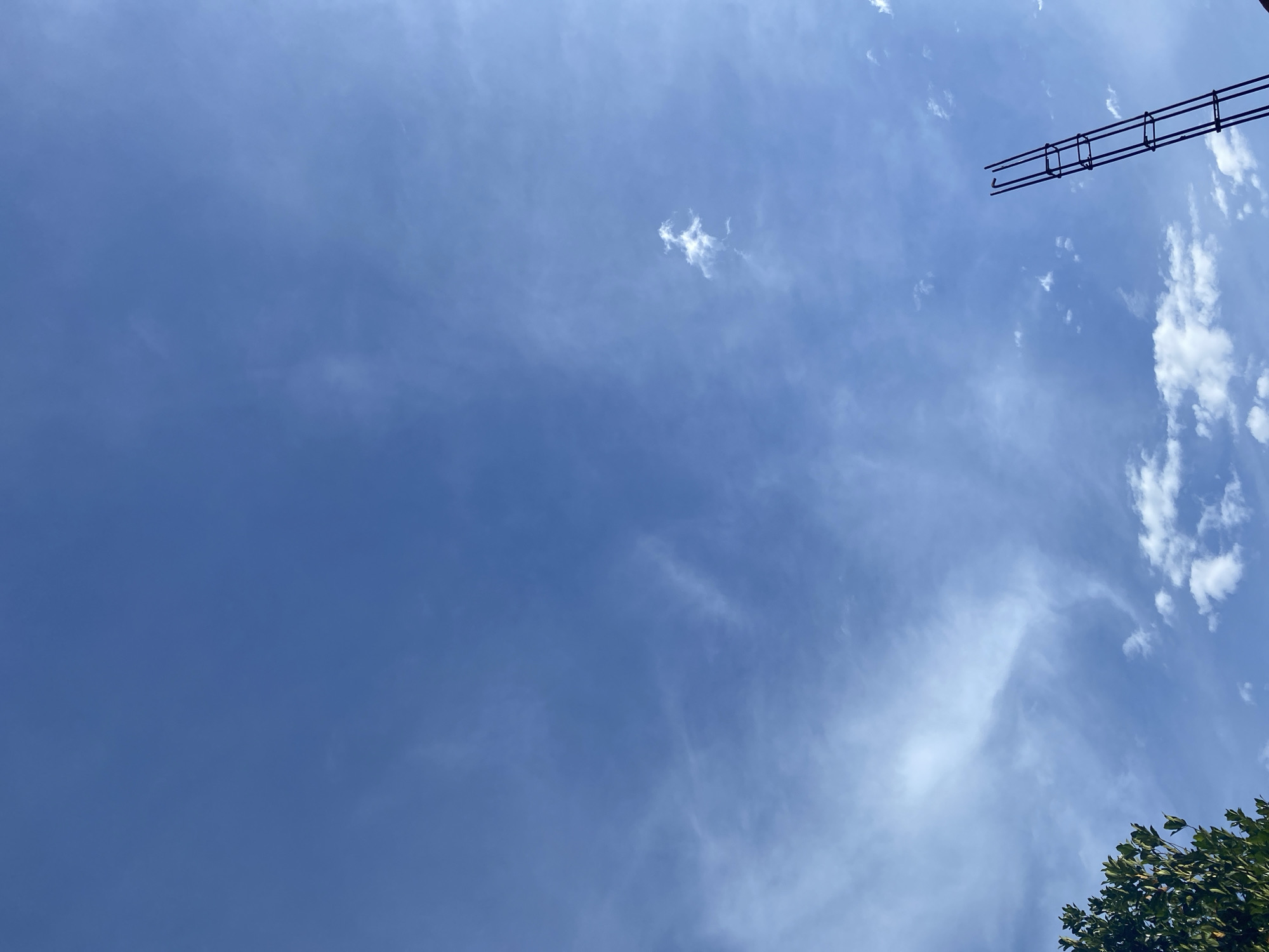 The image shows a clear blue sky with scattered white clouds, indicating fair weather. In the foreground, a portion of lush green foliage is visible on the left side, and on the right, a dark, skeletal structure, possibly rebar from a construction site, extends upwards into the sky. The lighting suggests it is daytime, with the sun likely positioned out of frame. There are no people or animals present in the image. The scene appears to be an outdoor environment, possibly in a developed area given the presence of the construction element. The location is identified as Banda Aceh, Indonesia.