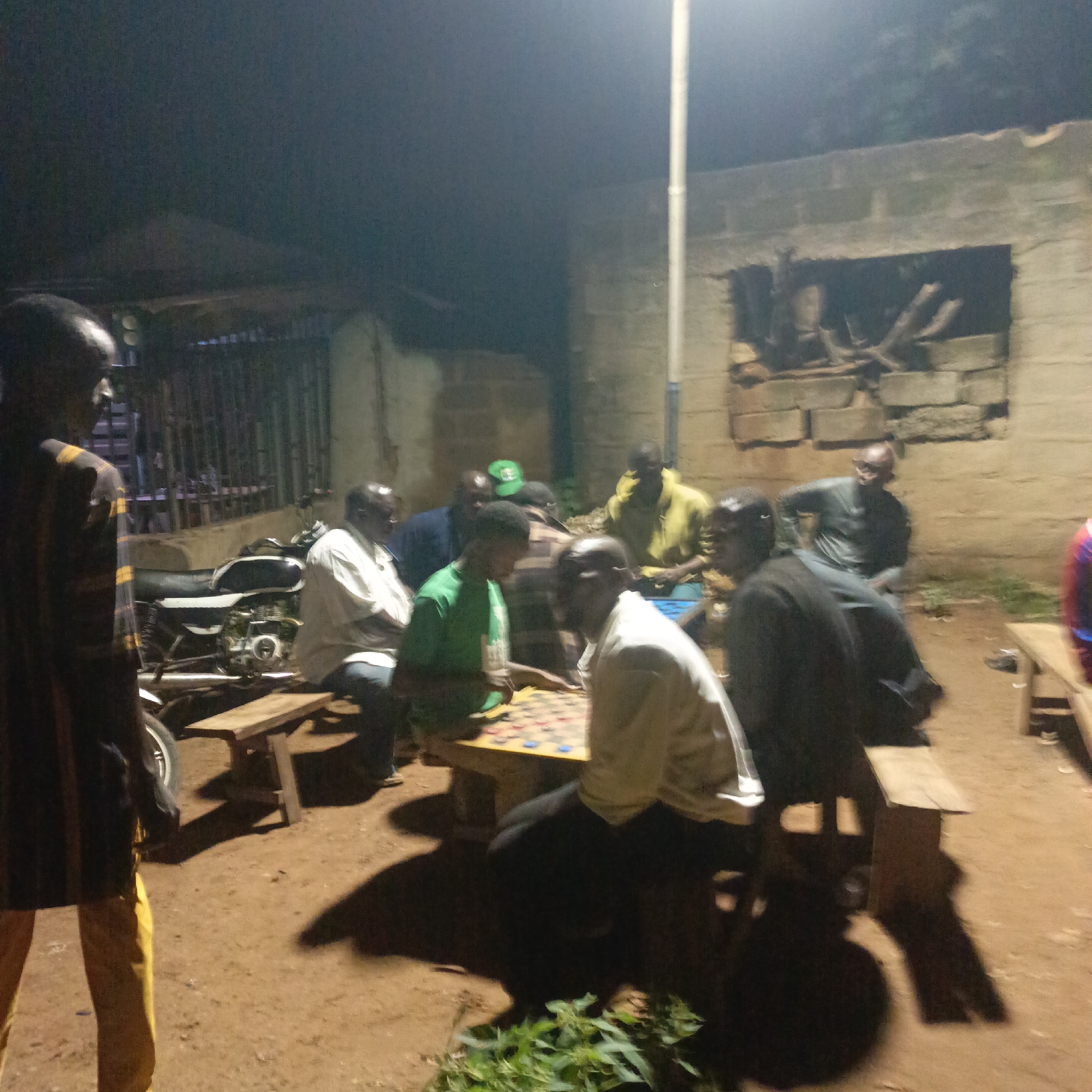 In Akwanga, Nigeria, a group of men gather outdoors at night for what appears to be a game. The scene is illuminated by artificial light, casting long shadows on the dirt ground. A motorcycle is parked to the left. In the center of the image, several men are seated around a low table, engaged in a game that involves placing circular pieces on a board with a grid. One man, wearing a green shirt, is actively moving pieces on the board. To the left, a man in a striped shirt and yellow trousers stands observing. Behind the players, a brick wall structure with an opening containing firewood is visible. The atmosphere suggests a casual social gathering in a semi-urban or rural setting.