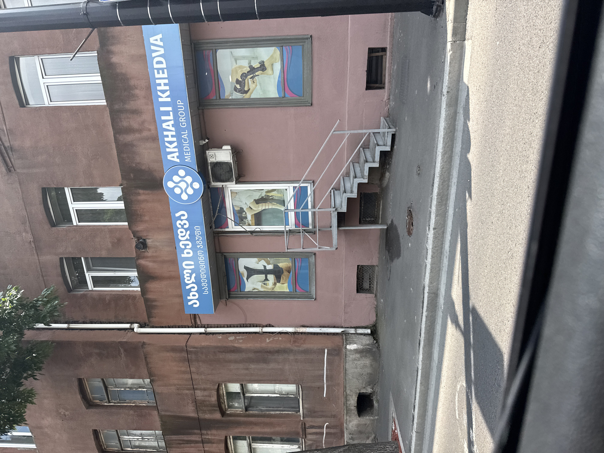 The image shows the exterior of a medical facility in Tbilisi, Georgia, identified by the signage "AKHALI KHEDVA MEDICAL GROUP." The building has a pinkish-brown facade with several windows. A blue sign above the entrance displays Georgian text and the English name of the medical group. Below the sign, there are two large display windows showcasing medical-related imagery, one depicting a person in a posture brace and the other a leg brace. An air conditioning unit is mounted on the wall above the entrance, and a metal staircase leads up to the entrance. The building is situated on a street with a sidewalk and road visible in the foreground. The overall environment appears to be an urban setting during daylight.