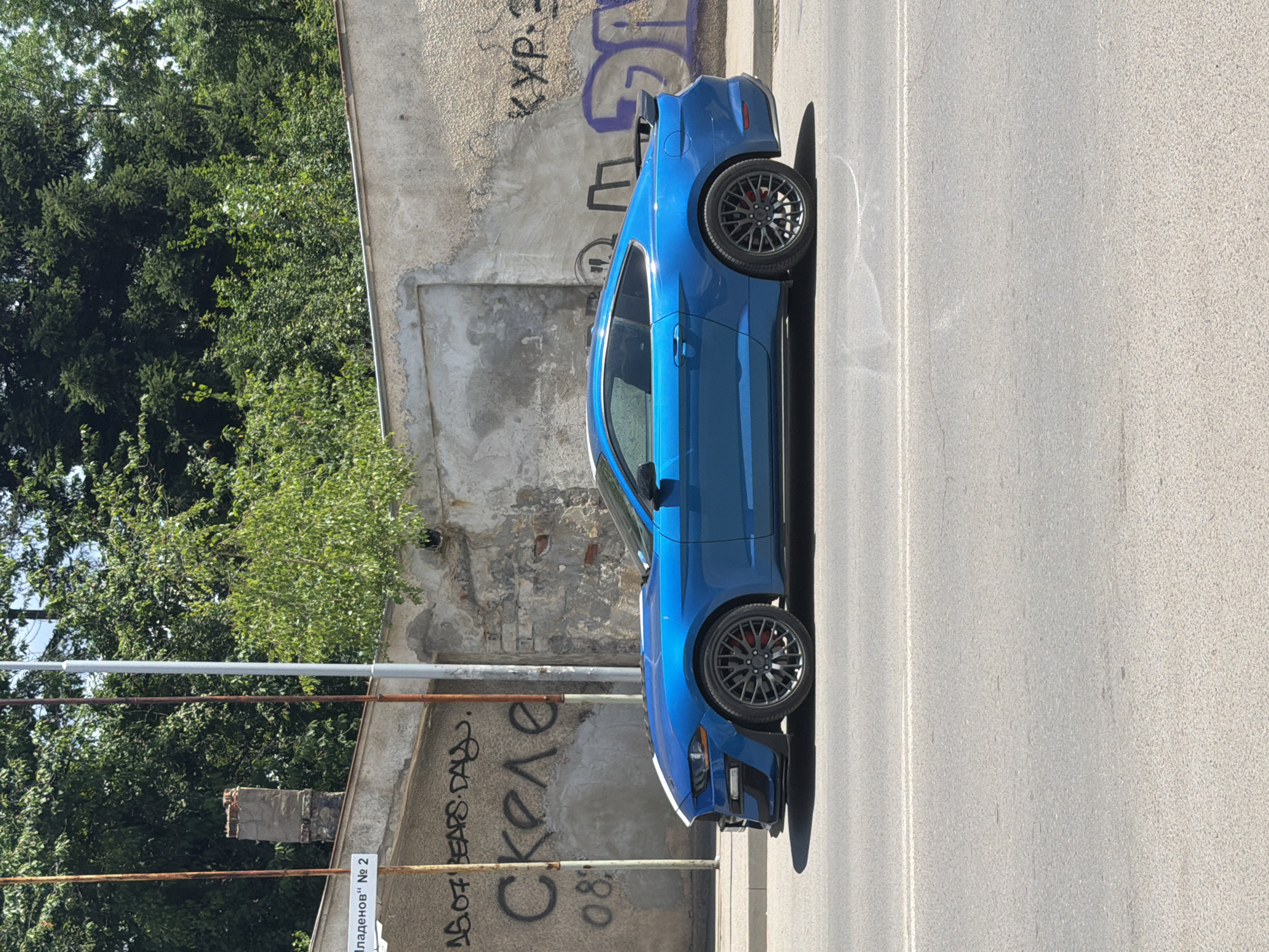 A bright blue Ford Mustang is parked on the side of a street in Sofia, Bulgaria. The car is positioned perpendicular to the viewer, its side facing the camera. The car has black multi-spoke rims with red brake calipers visible. The vehicle appears to be in good condition.
The scene is an urban setting with a weathered, graffiti-covered concrete wall on the left side of the image. The graffiti appears to be in Cyrillic script, with some letters and symbols visible. Behind the wall, lush green trees create a dense canopy. The street itself is paved with asphalt and has a white line marking a lane.
In the background, parts of a building are visible, including some brickwork and pipes. A small white sign with Cyrillic text is attached to the wall, which reads "Пладенов" No 2. The lighting suggests it is daytime, likely mid-morning or afternoon, with the sun casting a shadow from the car onto the asphalt. The weather appears to be clear and sunny. There are no people visible in the image. The overall impression is a snapshot of a car parked in a somewhat industrial or derelict urban area, contrasting with the vibrant green foliage.
