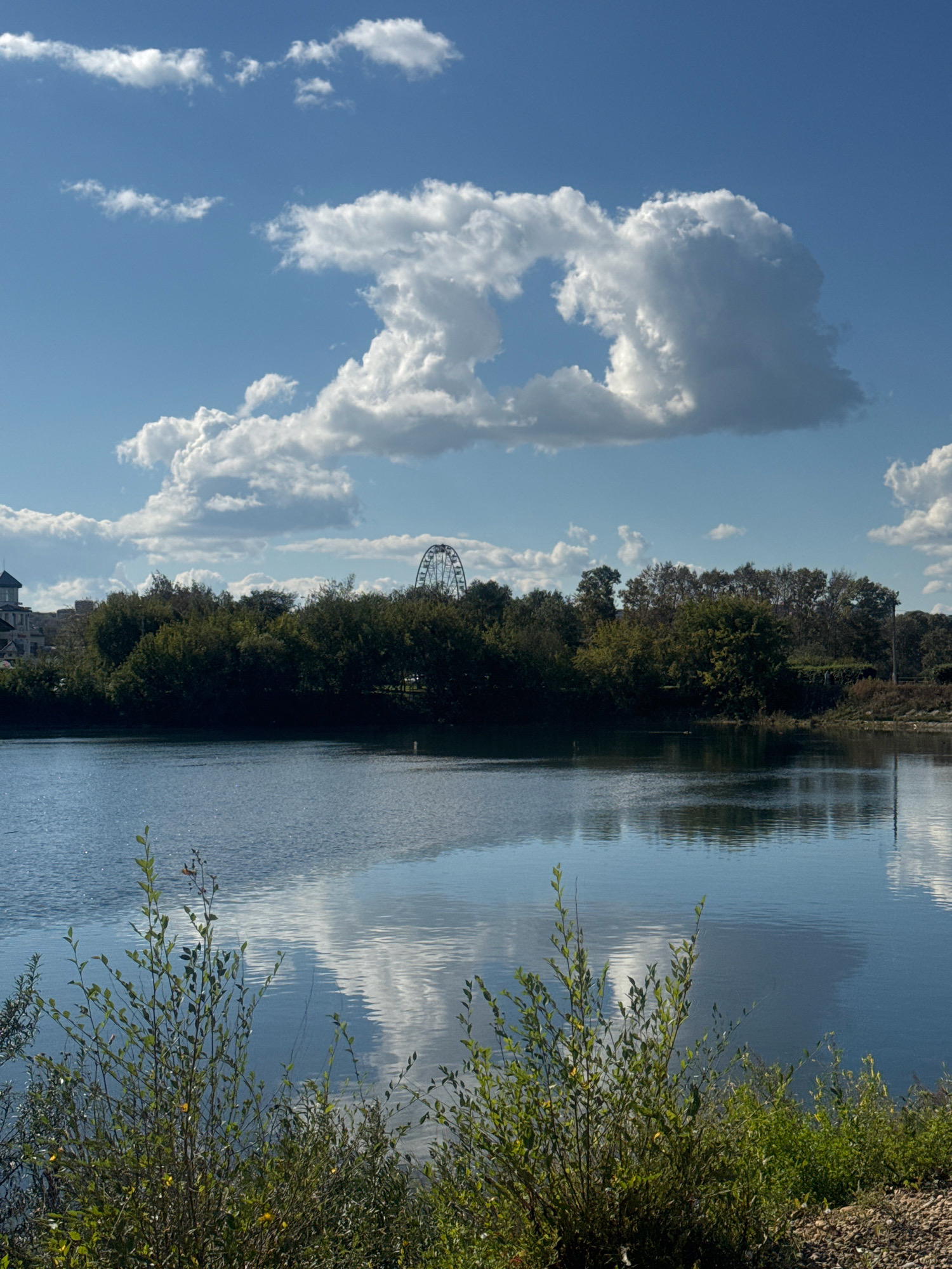 The image depicts a tranquil body of water with trees and a Ferris wheel in the background. The sky is a bright blue with fluffy white clouds casting shadows on the water. The foreground features green bushes and gravel. The reflection of the clouds and trees on the water adds to the serene atmosphere of the scene. There are no people or animals visible in the image. The Ferris wheel suggests the presence of an amusement park or a similar recreational area. The overall setting appears to be a park or a waterfront area on a clear, sunny day.
