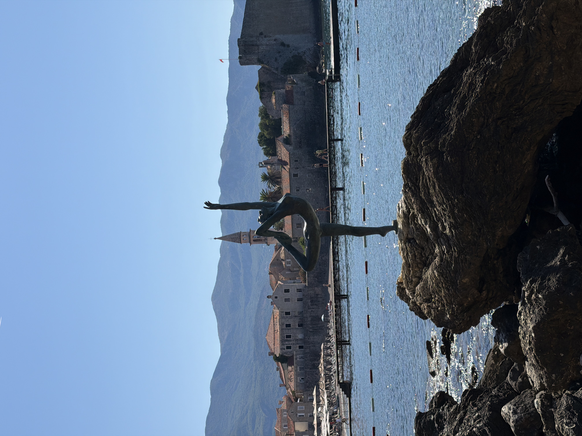A statue of a dancer is in the foreground, silhouetted against a bright blue sky and a body of water. The statue is positioned on a large rock. In the background, there is a beach with lounge chairs and umbrellas, and beyond that, a town with red-tiled roofs and a tall church steeple. A stone wall and a fort are also visible along the coast. The water is calm and sparkling in the sunlight. The overall scene suggests a sunny day in a coastal city.