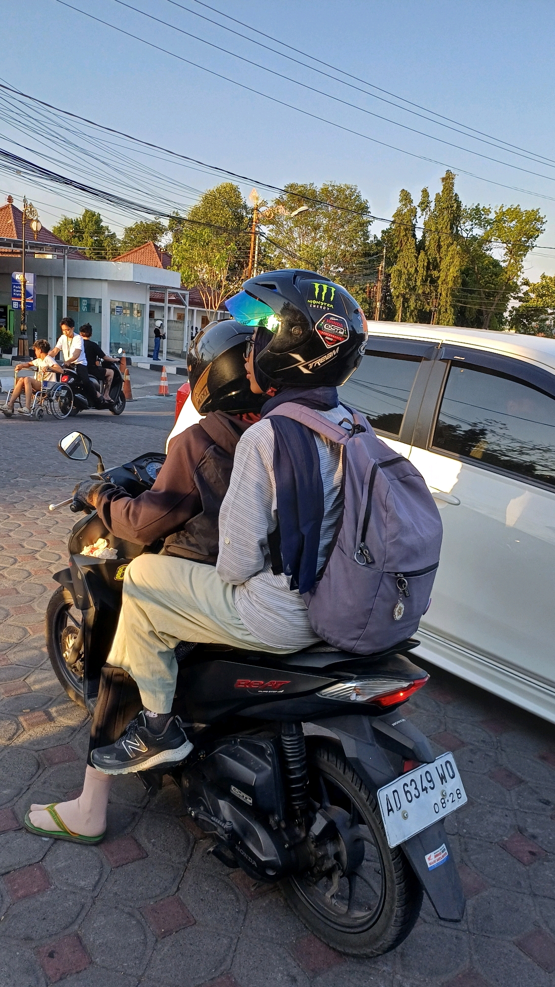 Two people are riding on a black motorcycle. The person in the front is wearing a black helmet with green accents and a blue visor. The person in the back is wearing a black helmet with the Monster Energy logo and other stickers. They are also wearing a light-colored striped shirt, a dark scarf, and a grey backpack. The motorcycle has a license plate that reads "AD 6349 WO 08-28".
In the background, there is a white car, a building with red-tiled roofs, trees, and power lines. To the left of the motorcycle, there is another motorcycle with two people on it, and a person in a wheelchair being pushed by another individual. This scene appears to be in the city of Klaten, Indonesia, based on the license plate format. The lighting suggests it is daytime, likely late afternoon or early evening due to the long shadows. The ground is paved with cobblestone-like material.