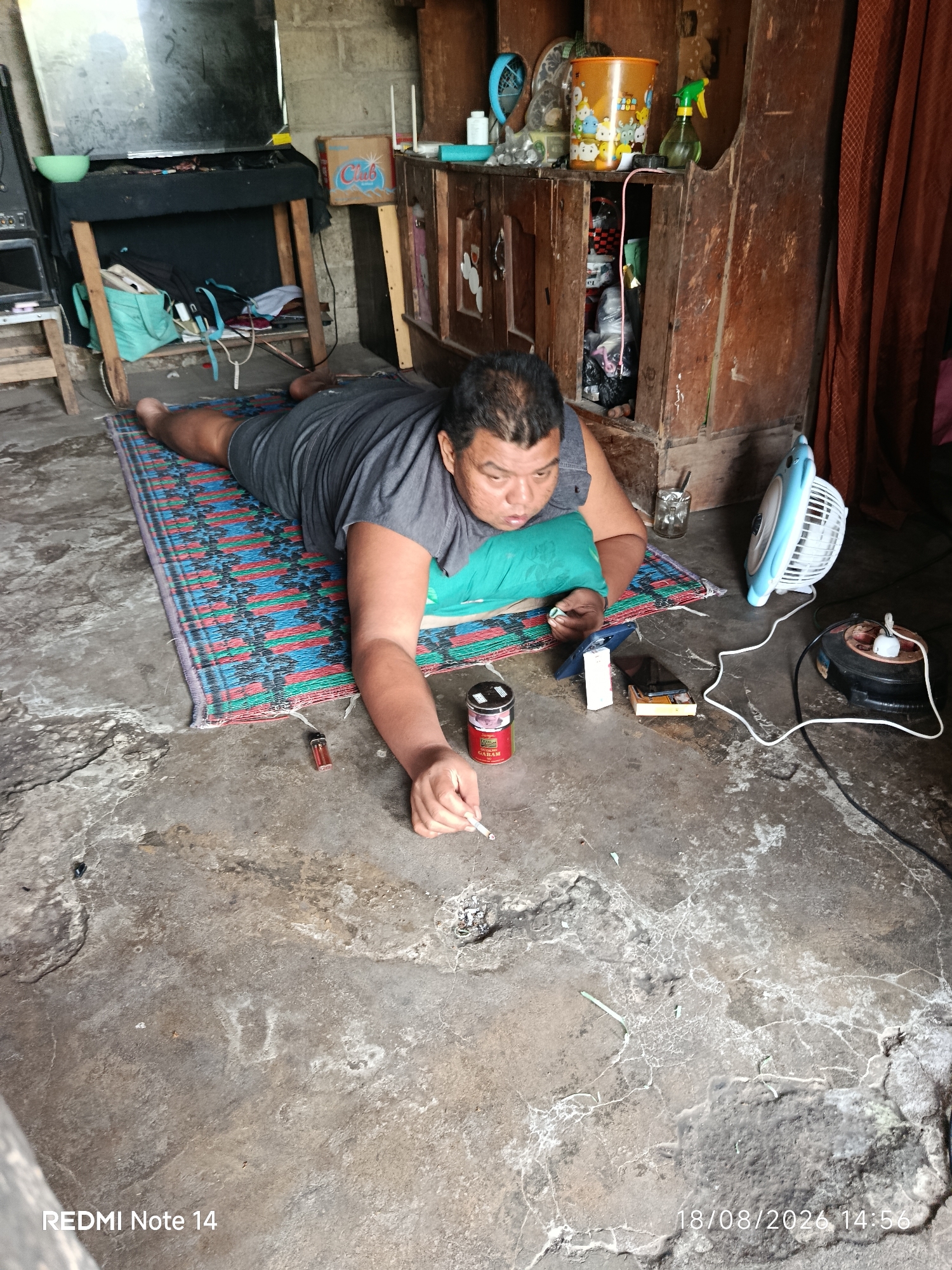 In a cluttered room with a concrete floor, a man is lying on his stomach on a colorful woven mat, wearing a grey t-shirt and shorts. He is holding a lit cigarette in his right hand and appears to be smoking. A green pillow is placed under his head. To his right, on the mat, is a red and silver cigarette pack. Nearby, on the floor, are a lighter, a smartphone, and a white rectangular object. Further to the right, a small blue electric fan is plugged into a power strip. The background reveals wooden shelves filled with various items, including a bucket, spray bottles, and a curtain. The timestamp "18/08/2026 14:56" and "REDMI Note 14" are visible in the bottom corners, indicating the image was taken in Panggungrejo, Indonesia.