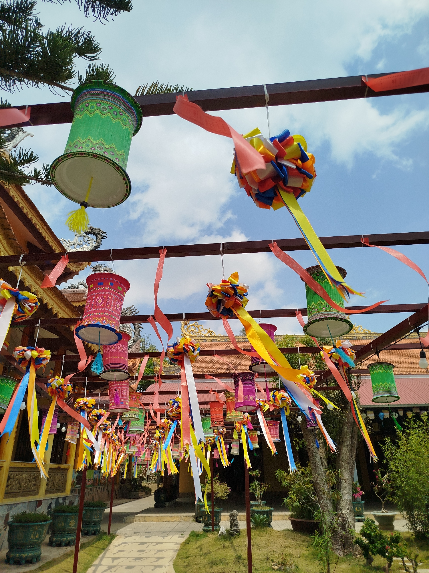 The image shows an outdoor space in Phát Chi, Vietnam, decorated with colorful lanterns and ribbon-adorned pom-poms suspended from a series of dark brown metal beams. The beams form a grid-like structure over a paved pathway leading through a courtyard. Green planters line the path, suggesting a manicured garden. To the left, a building with ornate architectural details and a gold-colored roof is visible, while trees and shrubs are seen on the right. The sky is blue with scattered clouds. The lanterns are cylindrical and decorated with patterns, and some have tassels hanging from the bottom. The pom-poms are multi-colored and feature long ribbons. The scene evokes a festive or celebratory atmosphere.
