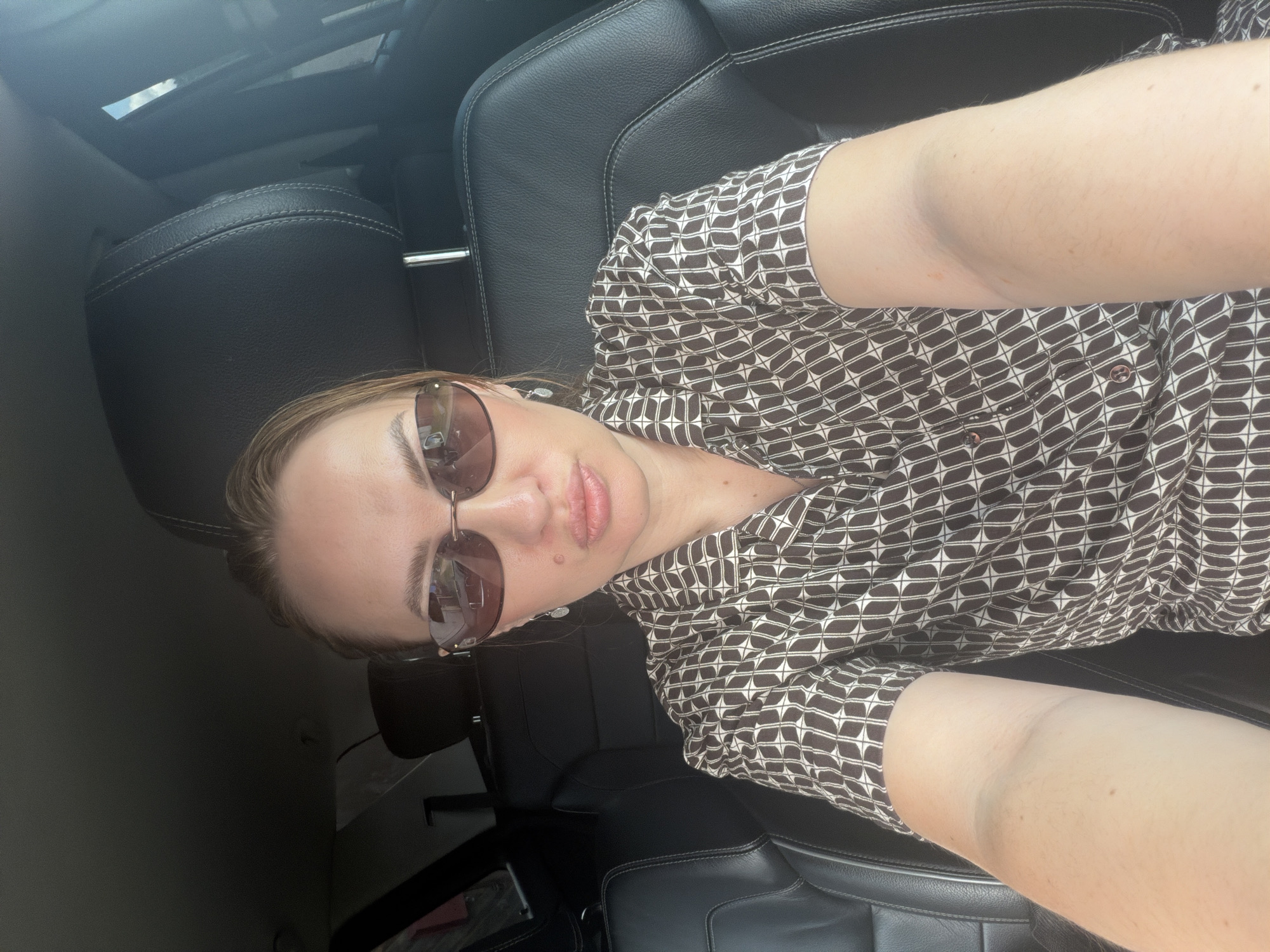 A woman is taking a selfie inside a car. She is wearing sunglasses and a patterned shirt. The interior of the car appears to be leather and the background is slightly out of focus. There is no information in the image to determine the location in Mezhevoy, Russia.
