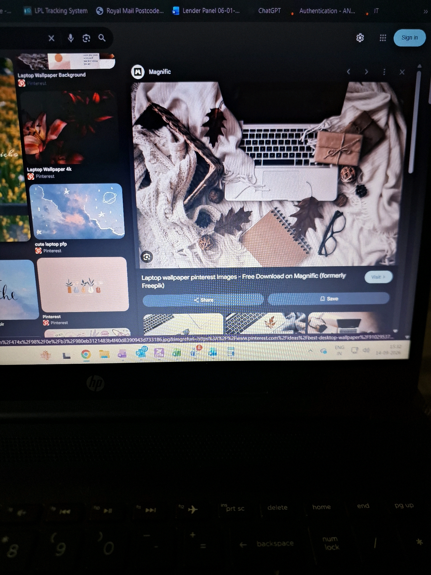 This is a close-up, high-angle, eye-level shot of a laptop screen displaying a Pinterest search for "Magnific" wallpaper. The screen shows a grid of images, with the main image being a cozy, autumnal flat lay of a laptop, blankets, autumn leaves, a notebook, and wrapped gifts. Below this main image are thumbnails of other related wallpapers, including floral designs and a celestial theme. The laptop itself appears to be an HP model, indicated by the logo visible on the keyboard. The screen also shows browser tabs at the top, with titles like "LPL Tracking System" and "Royal Mail Postcode." The bottom of the screen displays the taskbar with various icons and the time "15:32" and date "14-09-2026". The image is taken from a perspective above and slightly to the right of the laptop. The ambient lighting is dim, suggesting the photo was taken in a low-light environment. There is no information in the image to suggest it is located in Yamunanagar, India; the context appears to be a digital screen.