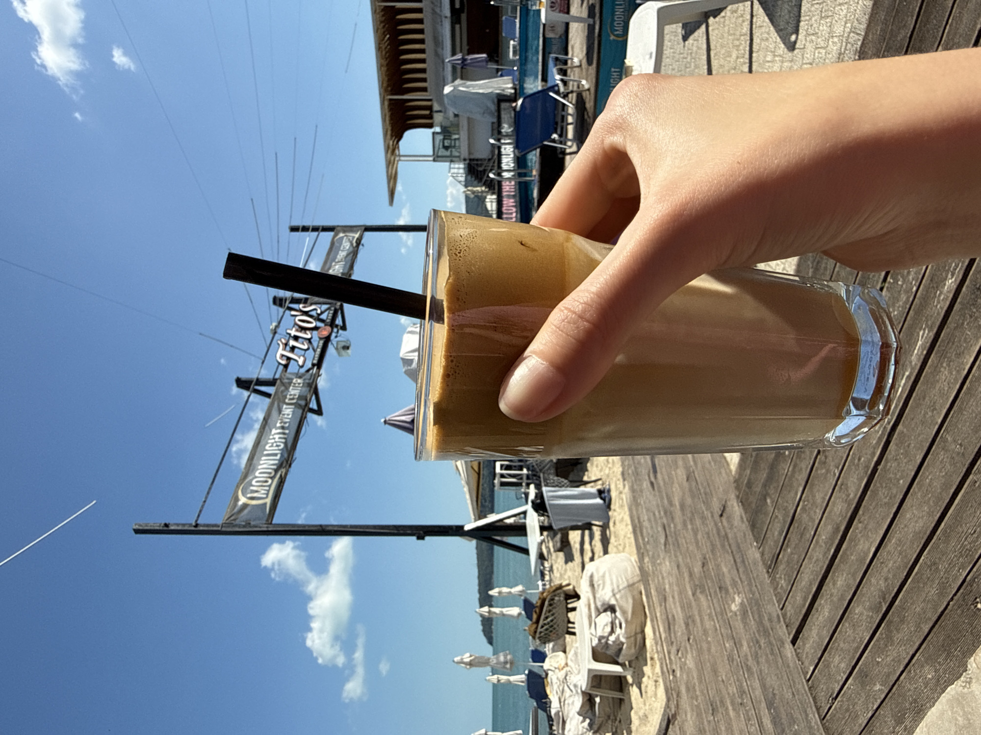 A hand holds a glass of iced coffee on a sunny day at Moonlight Event Center in Unknown, Unknown. The backdrop features a beach with white umbrellas, tables, chairs, and the sea. The sky is blue with scattered clouds. The wooden deck and nearby buildings are also visible.