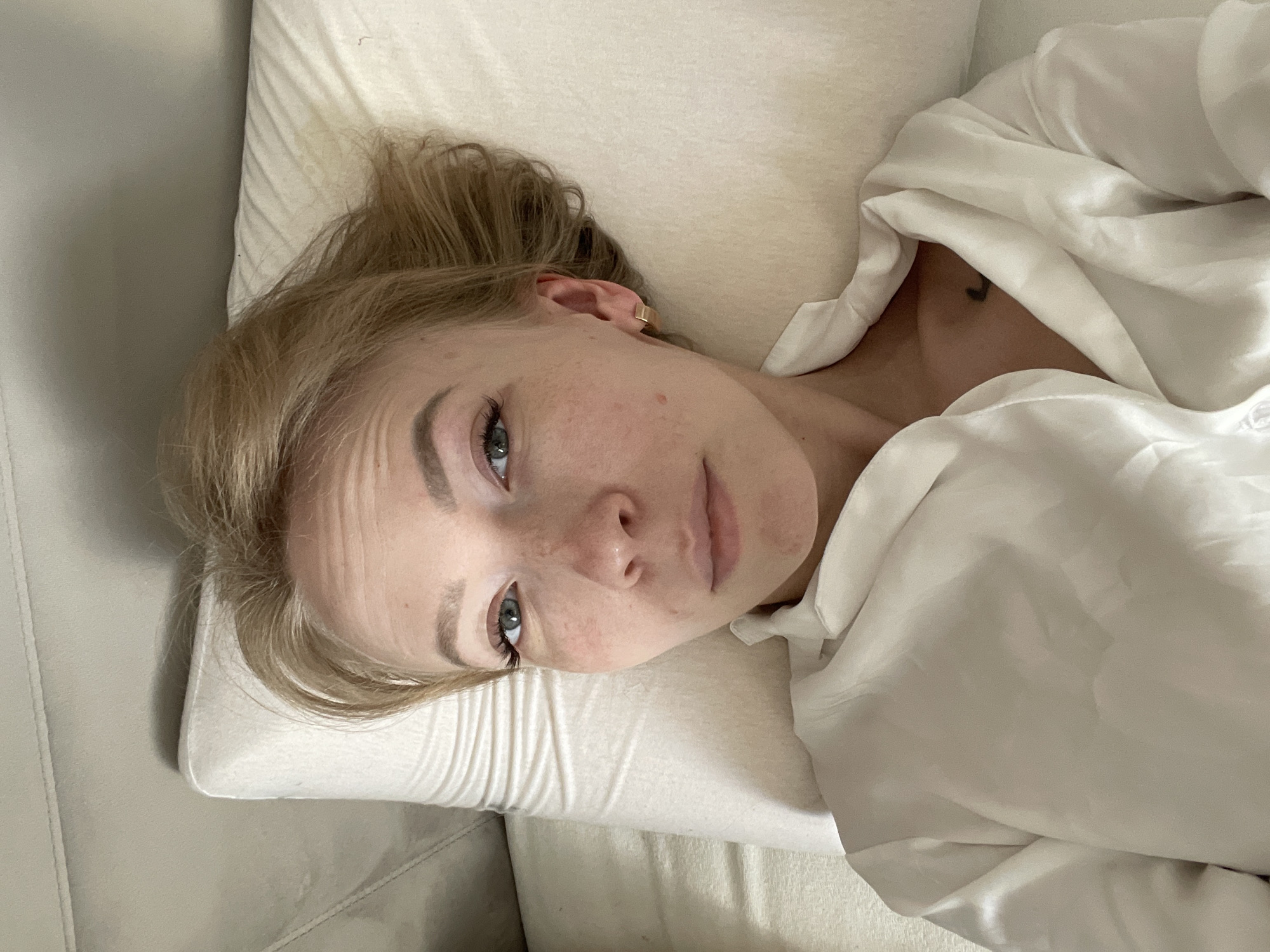 A light-skinned woman with blonde hair is lying down. She is resting her head on a white pillow, and part of a white shirt or robe covers her body. A portion of a light gray couch is visible on the left. A small tattoo is visible on the woman's chest. The photograph appears to be taken indoors, with natural lighting. There is no visible indication of Perm, Russia.