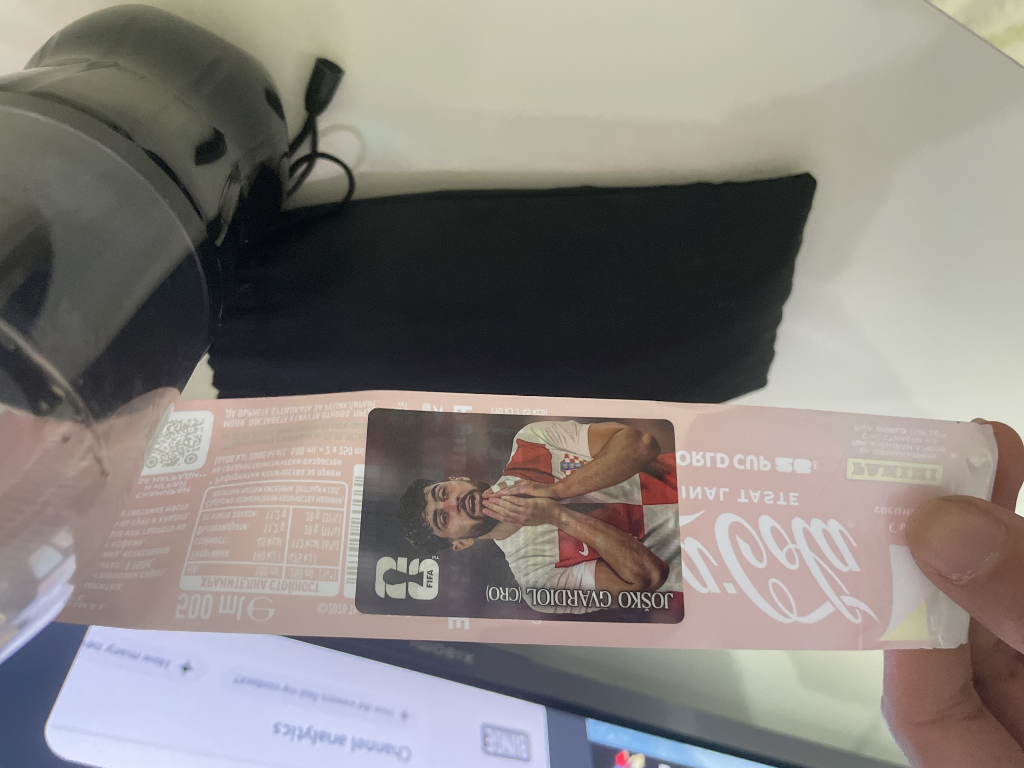 An indoor scene is captured from an overhead perspective, focusing on a person's hand holding a pinkish-red beverage wrapper or packaging. The central feature is a sticker on the wrapper depicting Croatian footballer Joško Gvardiol, wearing a white and red checkered national team jersey, with his hands clasped under his chin. The text "FIFA" and "JOŠKO GVARDIOL (CRO)" are clearly visible on the sticker.
The wrapper itself prominently displays the "Coca-Cola" logo (reversed due to the item's orientation) and the phrase "ORIGINAL TASTE". Other visible text on the packaging includes "200 ml" (reversed), a QR code, "SOSP", and various nutritional details. Several lines of text are in Cyrillic, such as "ОРИГИНАЛЕН ВКУС" (Original Taste) and numbers like "113 KCS".
In the upper left of the frame, a black, glossy object, possibly a speaker or part of an electronic device, is visible, along with a black power cord. Beneath it lies a black fabric item, such as a bag or cover. The upper background consists of a white surface, likely a desk. In the lower portion, a digital screen (from a laptop or monitor) is partially visible, showing text like "Channel analytics" and "BINTE". The setting appears to be a casual indoor environment, possibly in Ihtiman, Bulgaria, as suggested by the location context and Cyrillic script on the packaging.