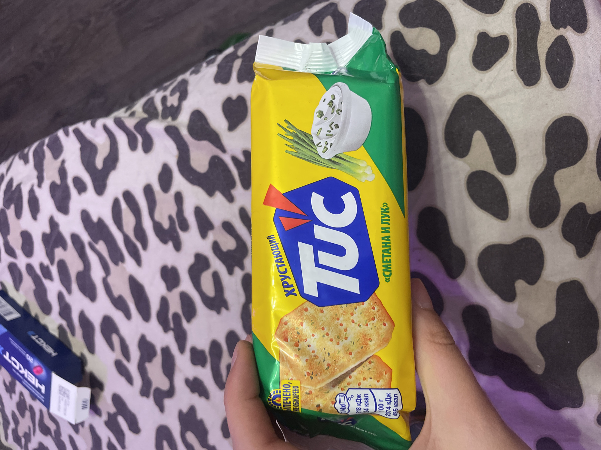 The image shows a hand holding a package of "TUC" crackers with a sour cream and onion flavor. The packaging is yellow and green with an image of the crackers and a bowl of sour cream with chives. The crackers themselves are thin and crispy, topped with herbs and spices. The background is a leopard-print fabric, possibly a blanket or bedspread. There is no information in the image or crops that provides any context about the city of Vaganovo, Russia.