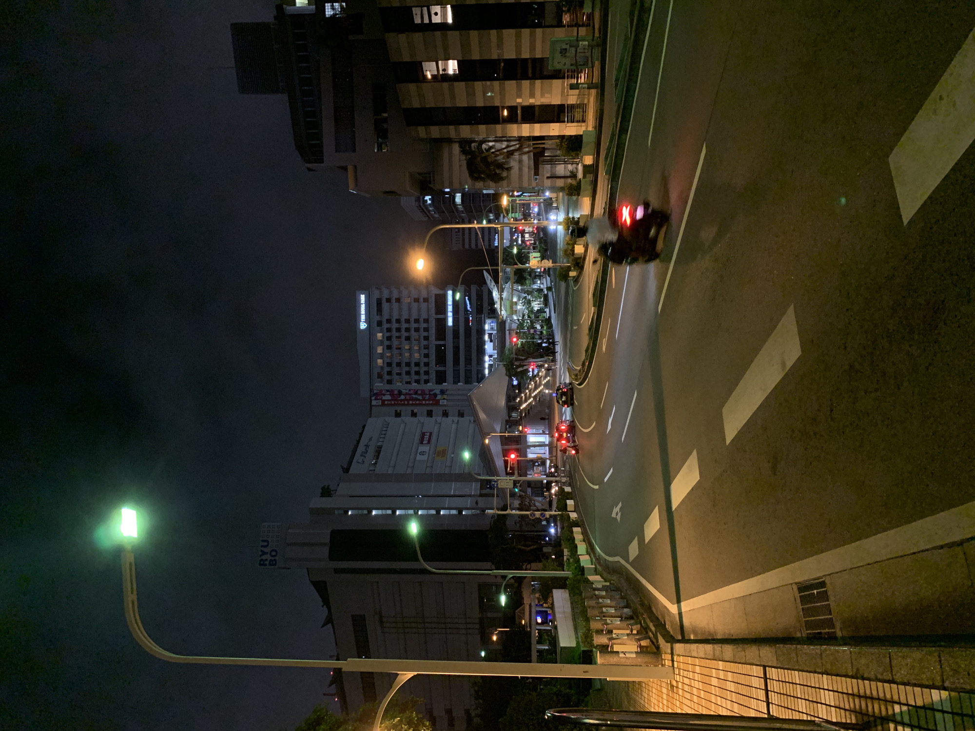 This is a night-time, eye-level, full shot of a street in Naha, Okinawa, Japan, likely taken near Kokusai Dori. The scene captures a curved road with two lanes of traffic, illuminated by streetlights and the lights of buildings. On the left side of the road, a sidewalk is visible with a pedestrian railing. The sidewalk is paved with checkered tiles, and a drainage grate is embedded in the curb. Several streetlights line the road, one of which is prominently displaying a green light.
In the background, modern buildings rise into the dark sky. One building on the left features a vertical sign with Japanese characters and "RYU BO" in English. Another building in the distance has a sign that reads "THE ROYAL PARK." Traffic lights are visible, with one showing a red light.
The road itself has clear white lane markings. A scooter with a rider is moving away from the camera on the right lane, its taillight illuminated red. Other vehicles are present in the distance, their headlights and taillights creating streaks of light. The overall atmosphere is urban and active, despite the late hour. The sky is dark with some diffused light, suggesting a slightly cloudy night.