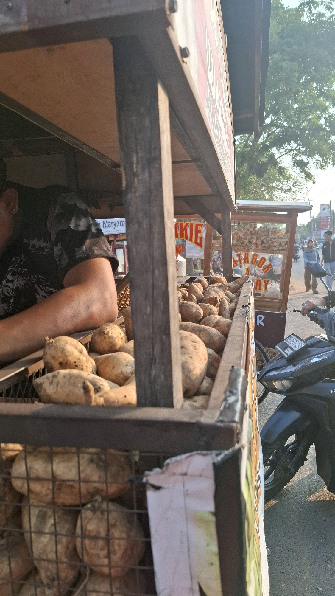 The image depicts a street food stall in Bandar Lampung, Indonesia, specializing in sweet potatoes. In the foreground, a large quantity of sweet potatoes is visible, piled in wire baskets. A person, wearing a black patterned t-shirt, is partially visible on the left, appearing to be tending to the stall. The stall itself is constructed of wood and metal, with signage indicating it sells "GORENGAN" (fried snacks) and "SINGKONG MARYAM" (a type of cassava). In the background, another food cart is visible with a sign that reads "GORENGAN." A motorbike is parked to the right of the frame. Several people are walking in the background, and trees and a street light are visible, suggesting an outdoor urban setting. The overall scene is consistent with a bustling street market or food vendor area in Indonesia.