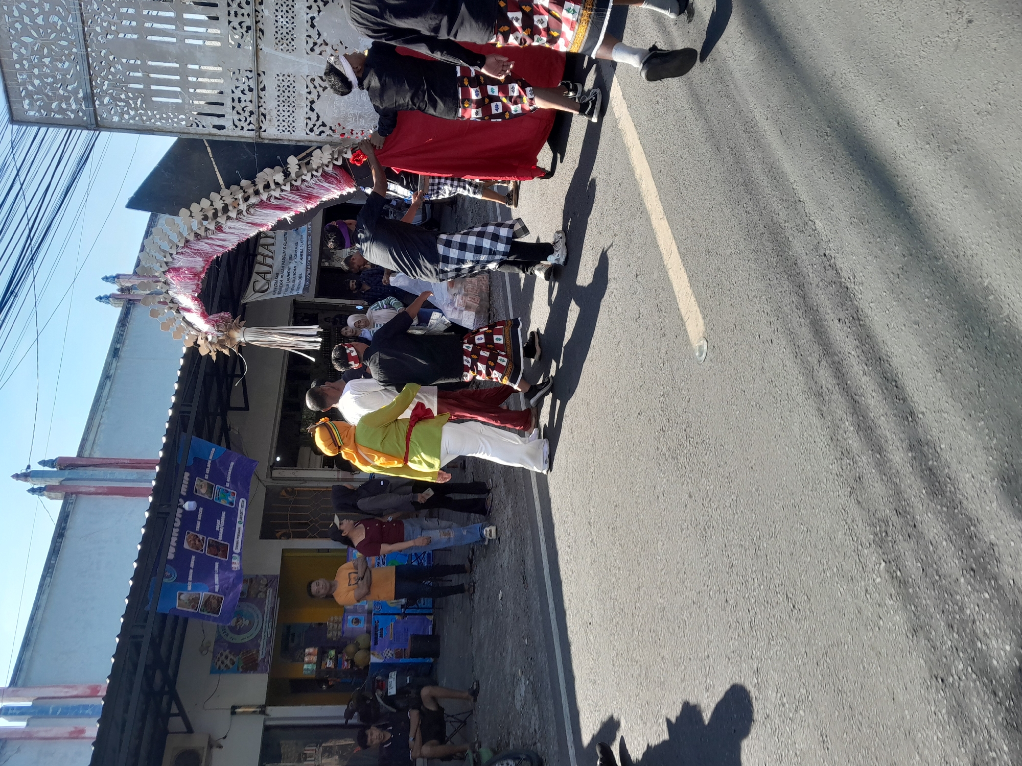 In Gentengwetan, Indonesia, a procession moves down a paved street under a clear blue sky. The procession is led by individuals adorned in traditional Balinese attire, featuring black shirts, checkered sarongs, and headbands. They are carrying a large, ornate archway constructed from white and pink paper or fabric. Behind them, others in similar ceremonial dress participate, some holding a large red cloth. Onlookers stand on the side of the road, observing the event. The background features a row of buildings, including shops with awnings and signage, suggesting a commercial area. The street is marked with a yellow line and shows shadows cast by the sun.