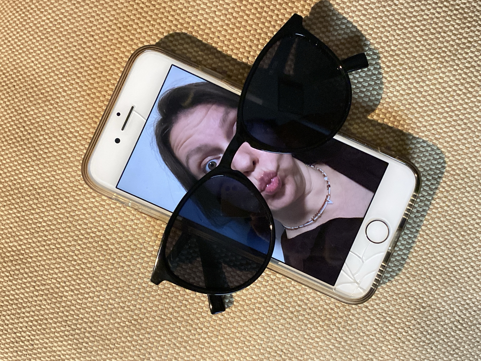 The image shows a white smartphone lying on a textured tan surface. The screen displays a selfie of a person with pursed lips wearing a necklace. Black sunglasses are placed diagonally across the phone. The bottom corner of the phone case is cracked. The location is unknown, and there is no verifiable information to indicate that the image was taken in Chaikovsky, Russia.